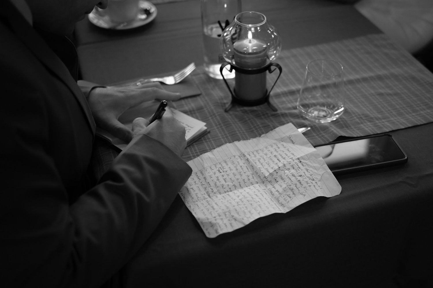 Black and white image of a person writing vows on paper at a candlelit wedding reception table.