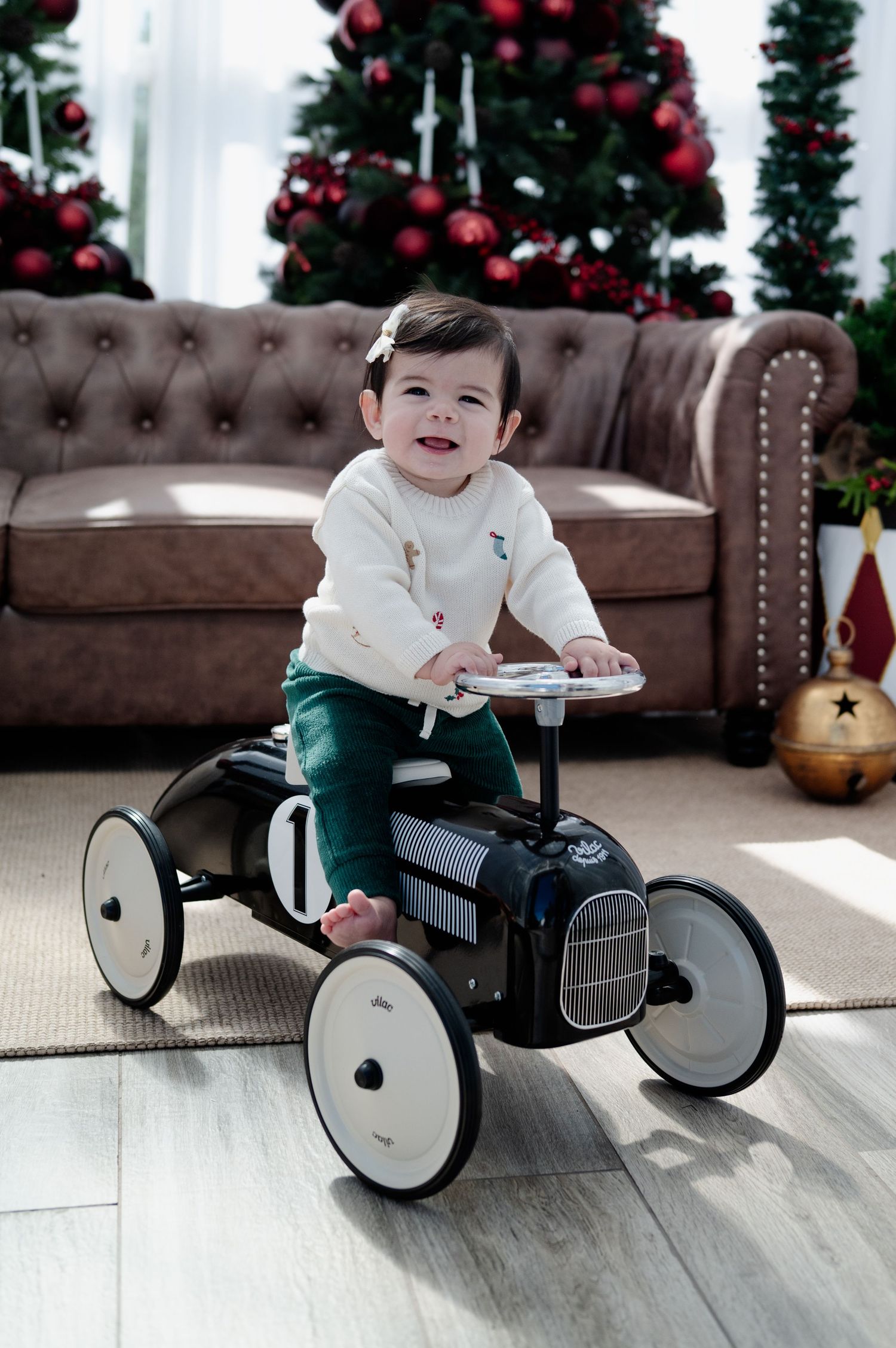 Smiling baby riding a vintage black toy car indoors with a decorated Christmas tree and tufted sofa in background.