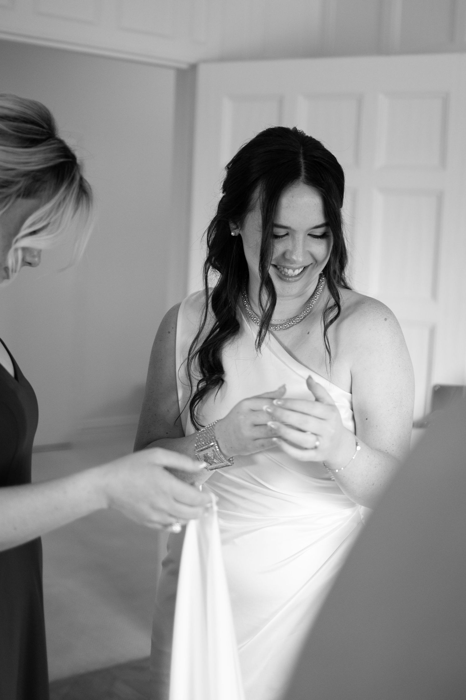 Bride smiling joyfully in white gown while bridesmaid helps with dress in black and white at The Pendry Hotel in Chicago Illinois.