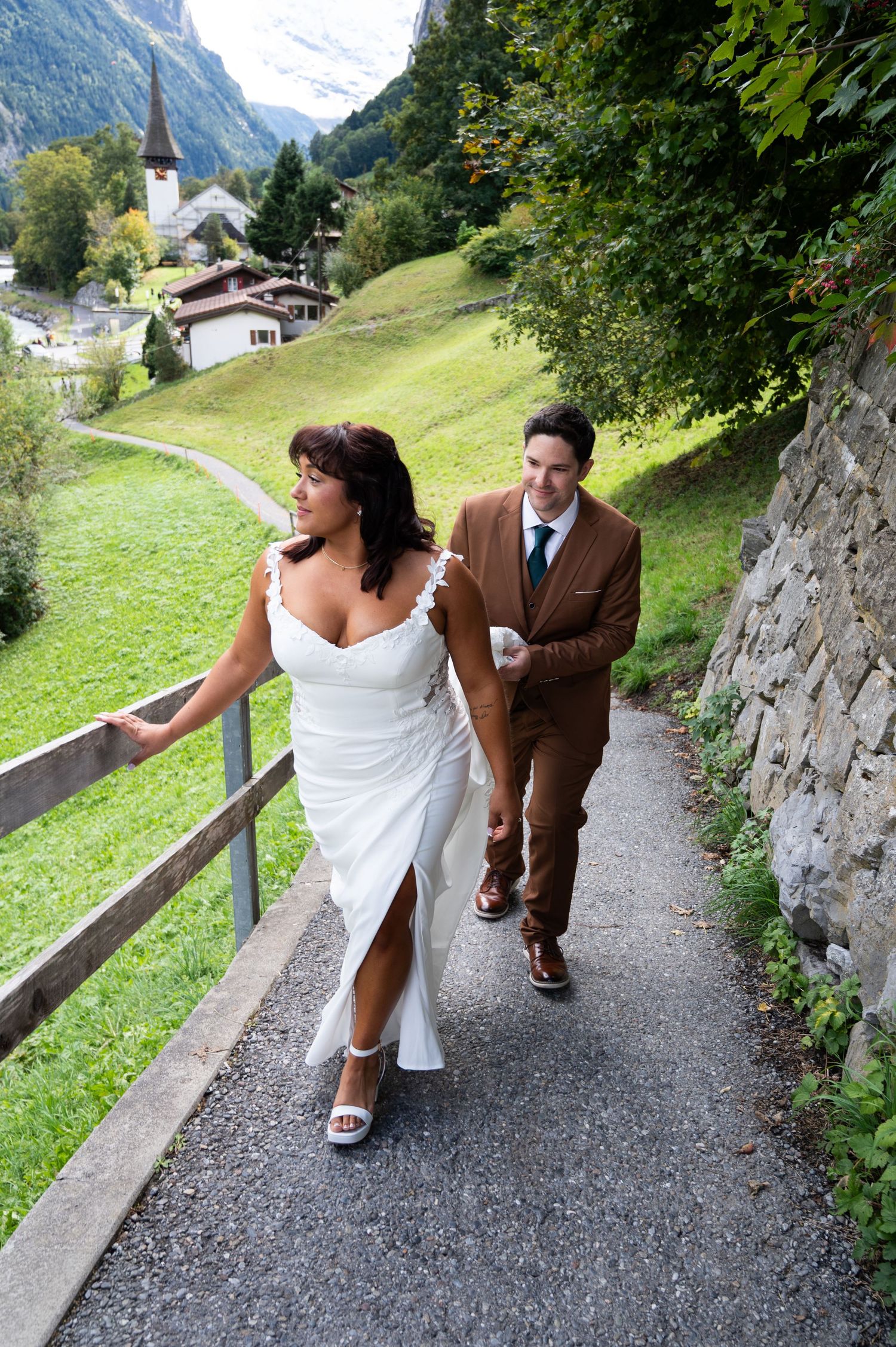 Happy bride and groom walk hand in hand along scenic Swiss village pathway on wedding day.