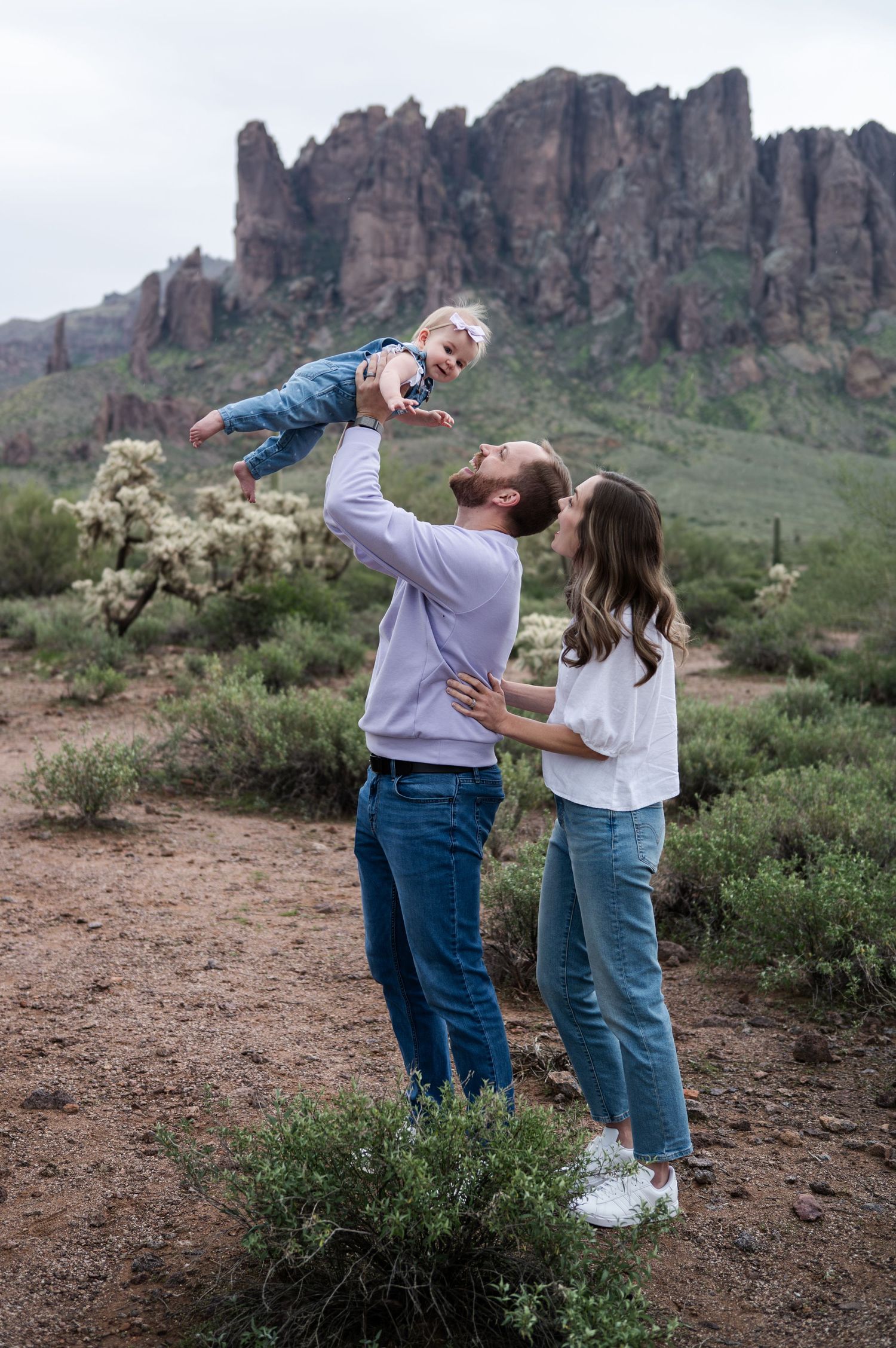 Family of three with toddler lifted high in desert landscape with rocky mountain backdrop, at the lost dutchman state part, arizona