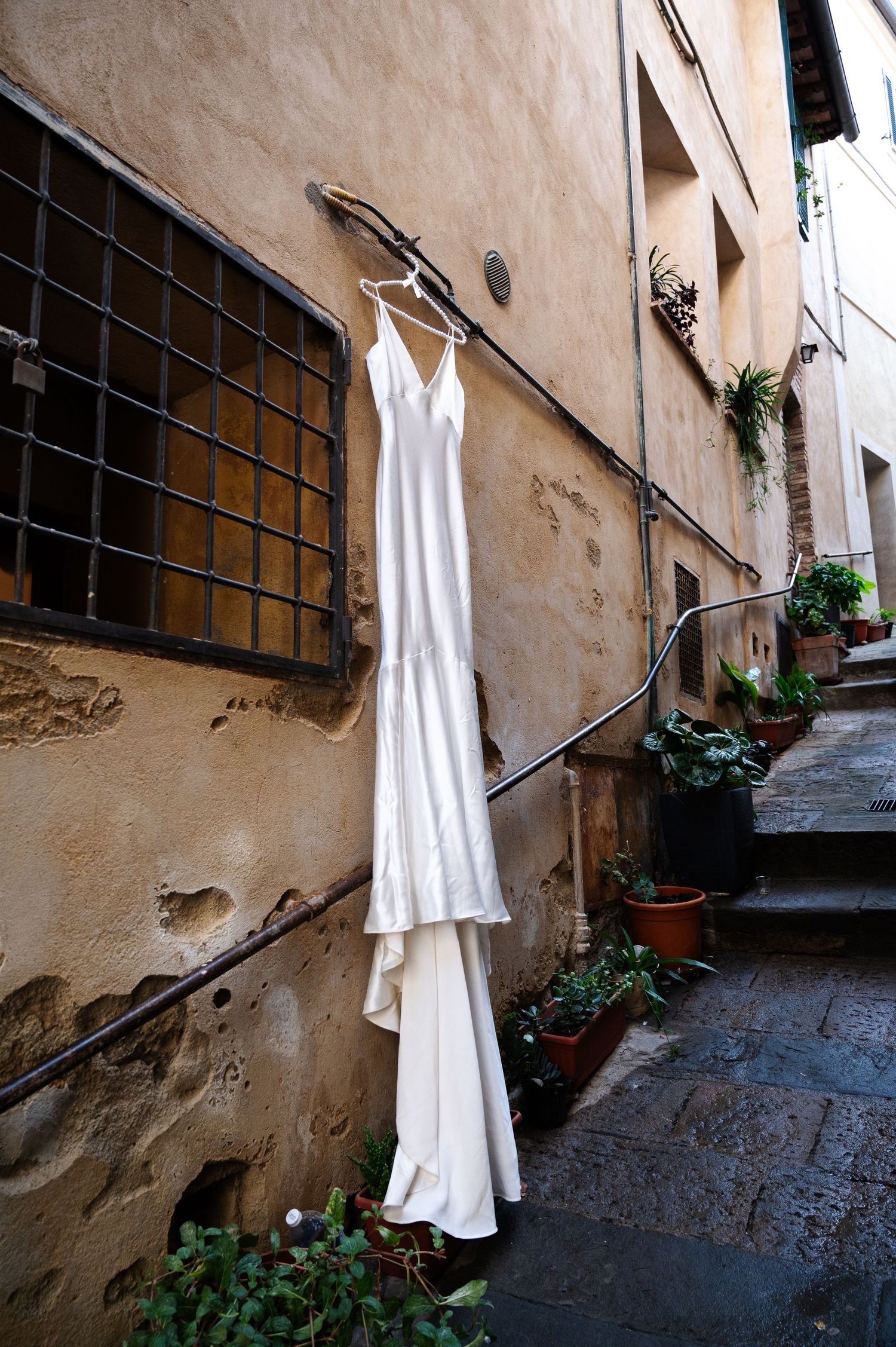 Elegant white satin wedding dress hanging on rustic Italian alleyway wall with stone steps and plants in Tuscany Italy.