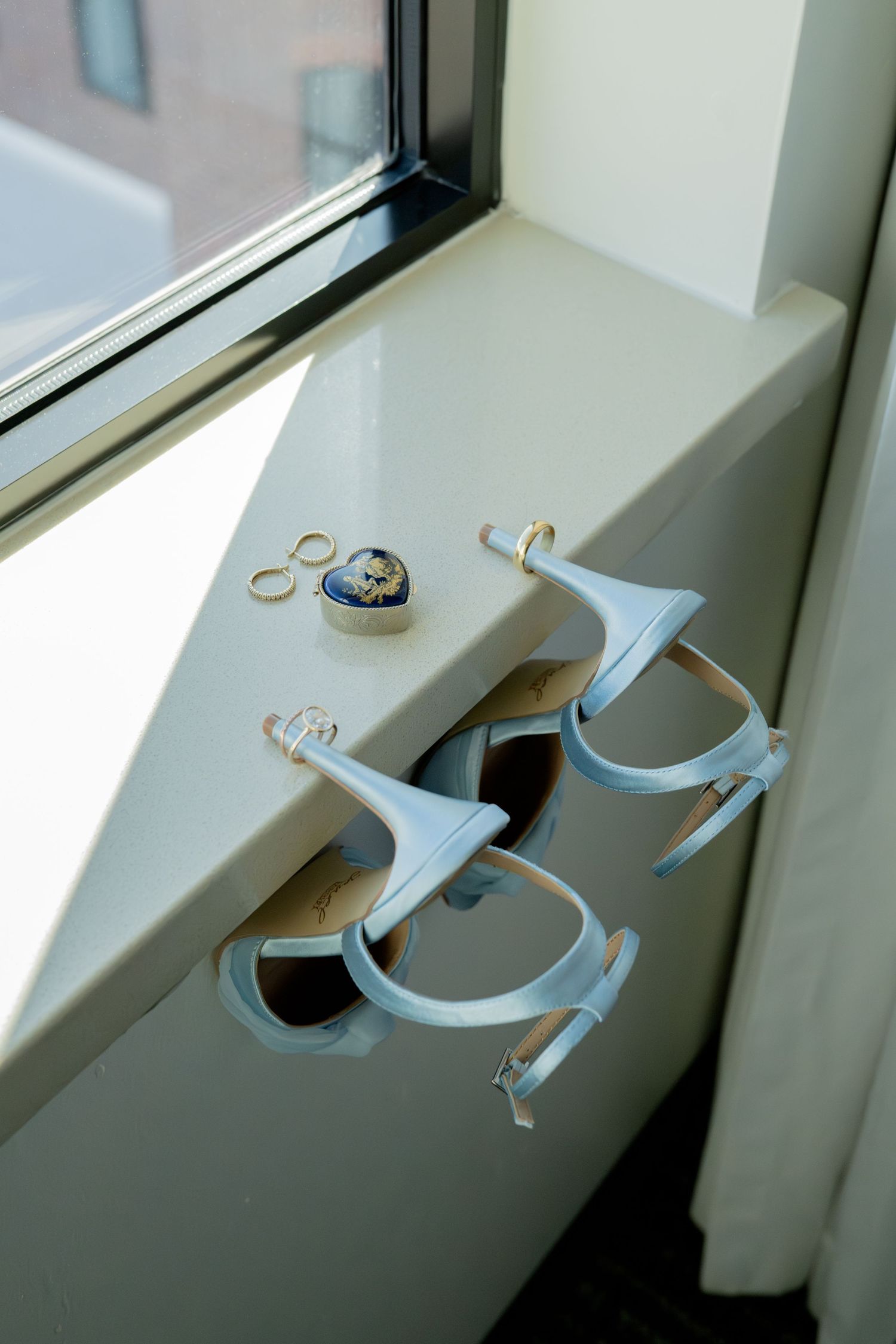Blue wedding shoes and gold rings on a sunlit windowsill with a small heart-shaped jewelry box.