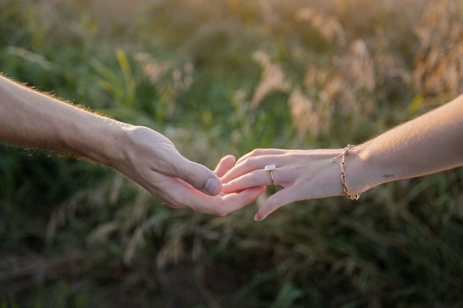 Two hands gently touching fingertips in a romantic moment outdoors, with a gold bracelet on the woman's wrist.