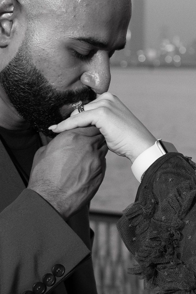 Black and white photo of a bearded man in a suit kissing a woman's hand with a diamond ring.
