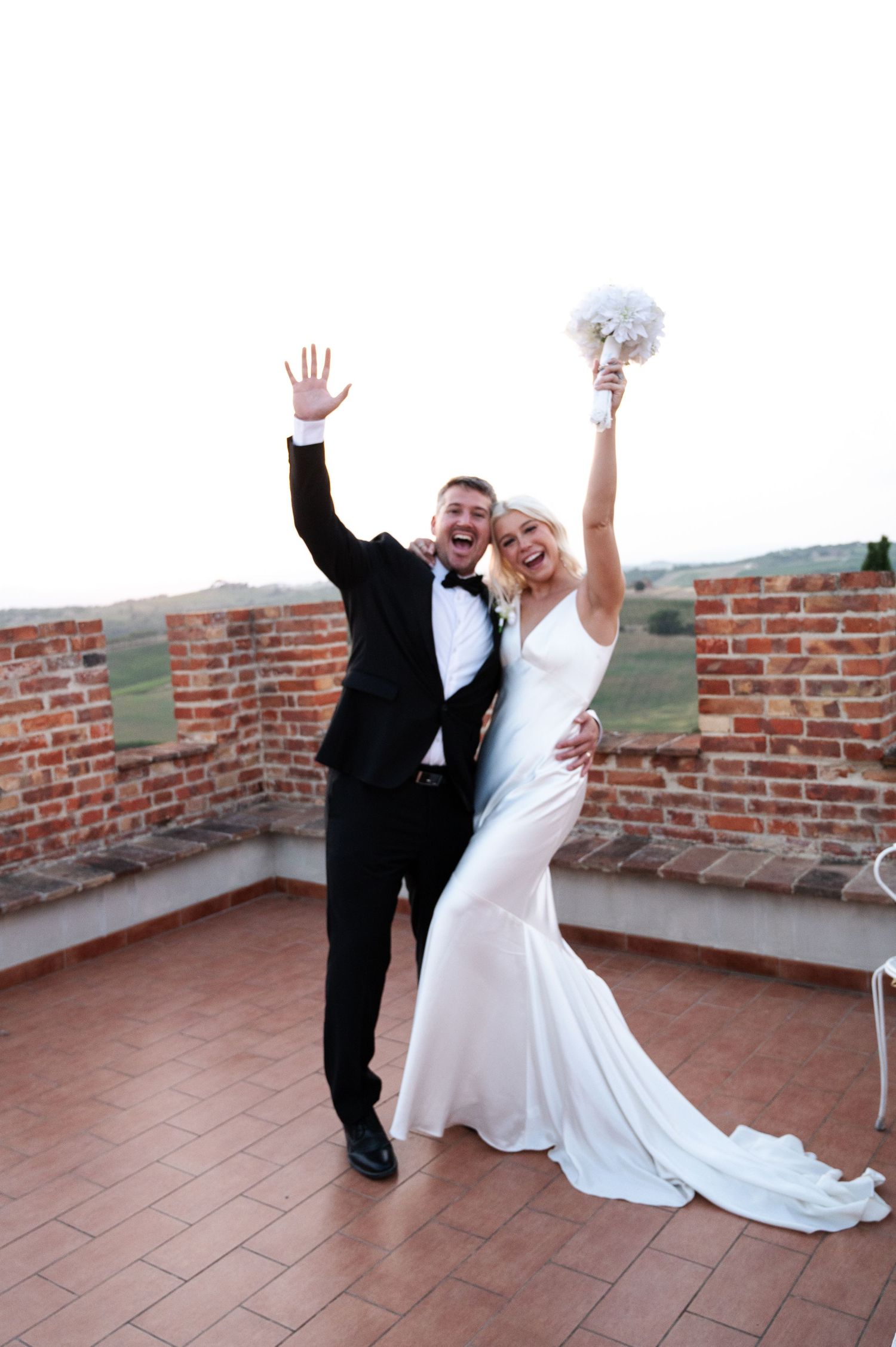 Joyful bride and groom celebrating on brick rooftop terrace, raising hands with bouquet at Italian wedding in Tuscany Italy.