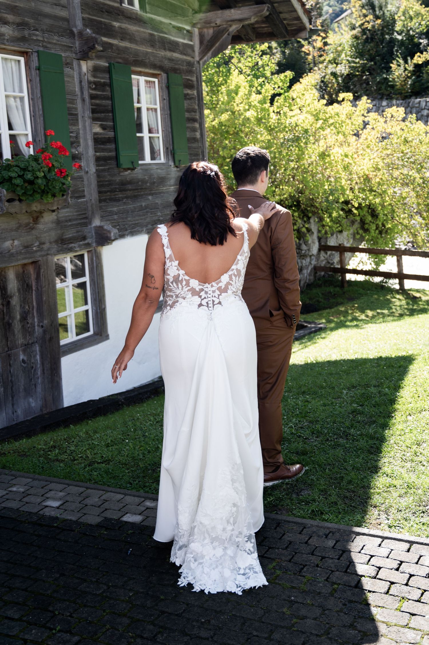 Bride and groom walking hand in hand toward a rustic wooden chalet with green shutters and flowers in Switzerland.