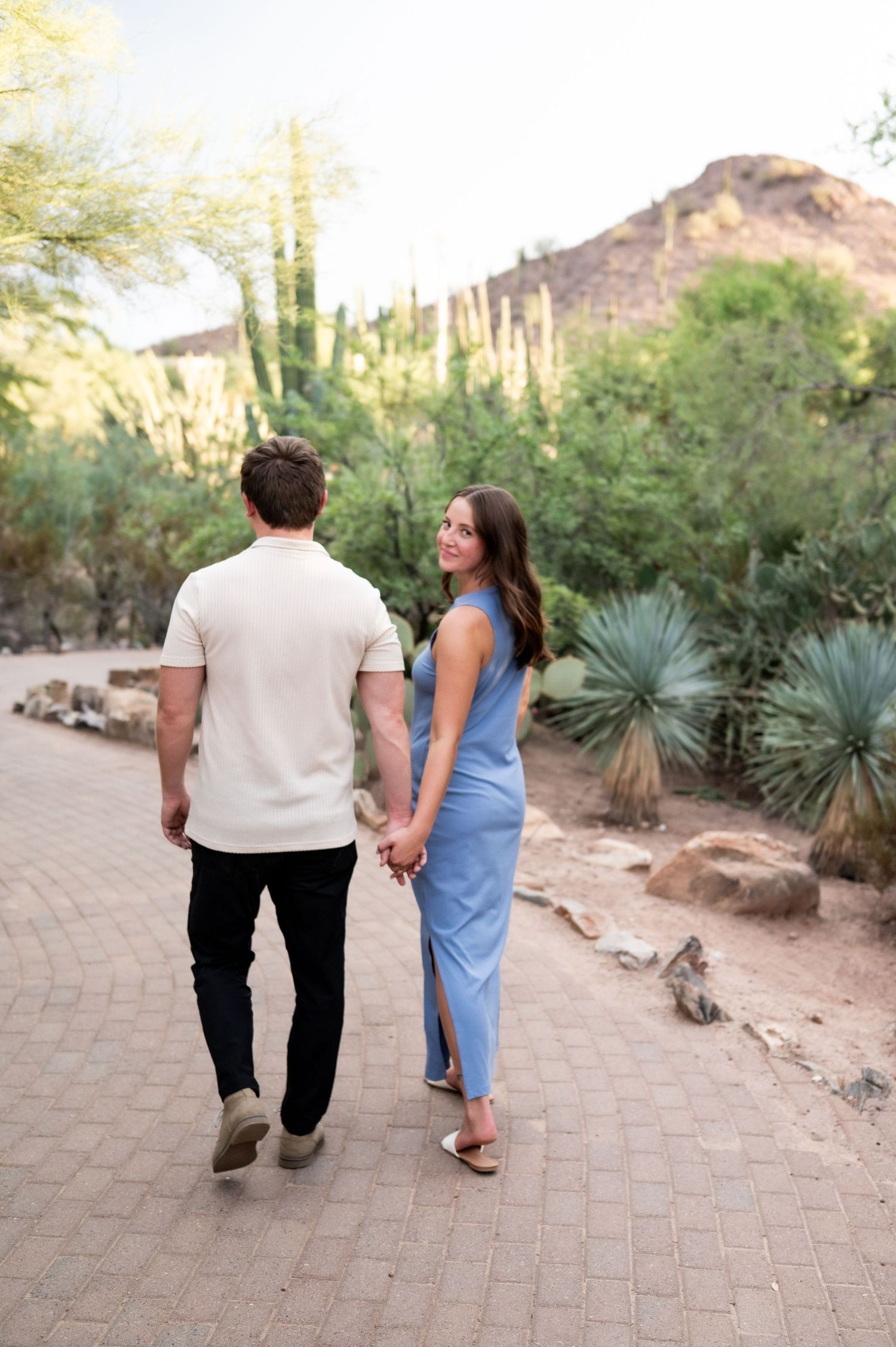 A couple holds hands walking on a brick path surrounded by desert cacti and mountains in a sunny outdoor setting.