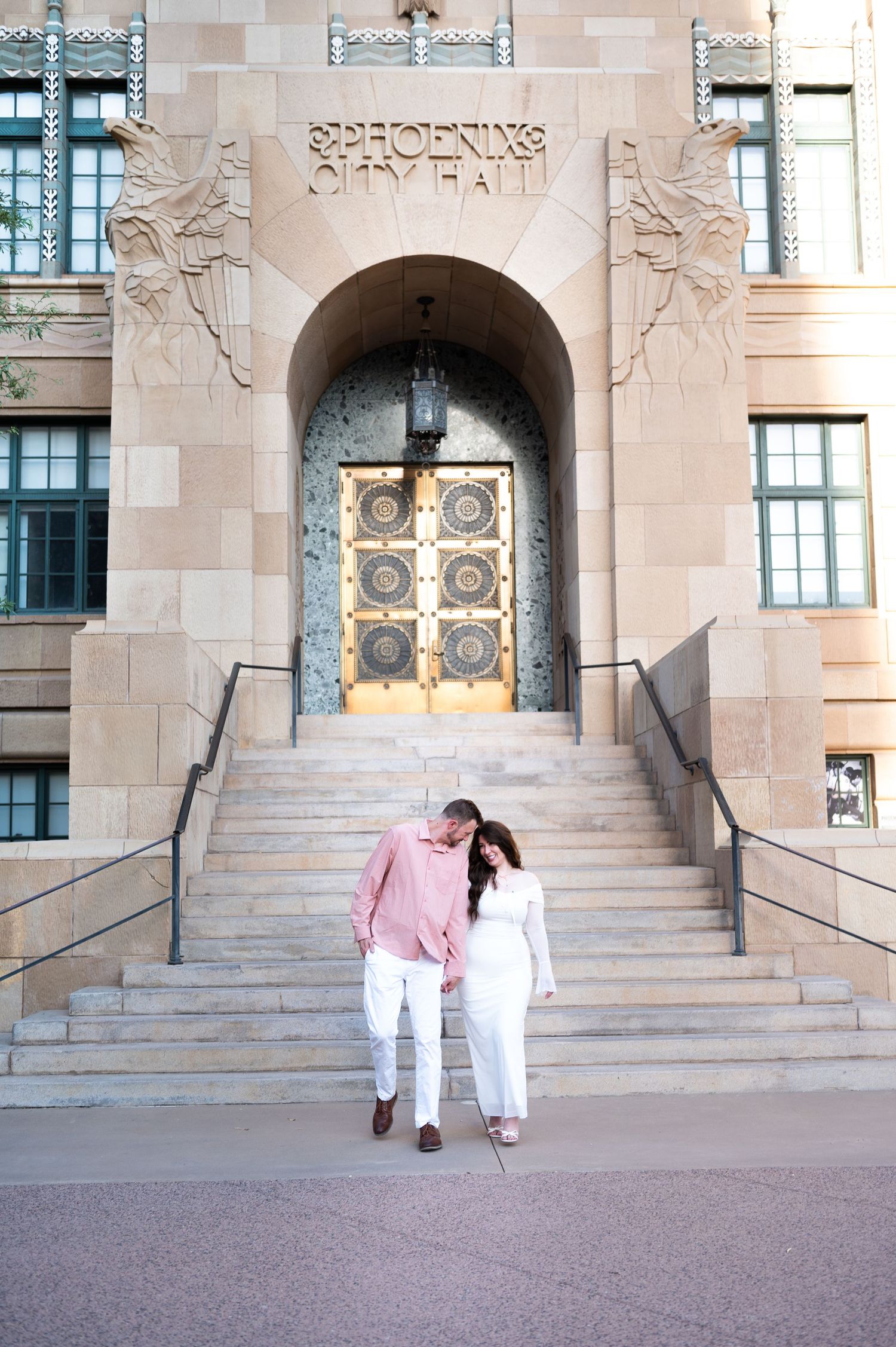 Romantic couple leans together on steps of Phoenix City Hall with ornate gold entrance doors at Old Courthouse in Phoenix Arizona.
