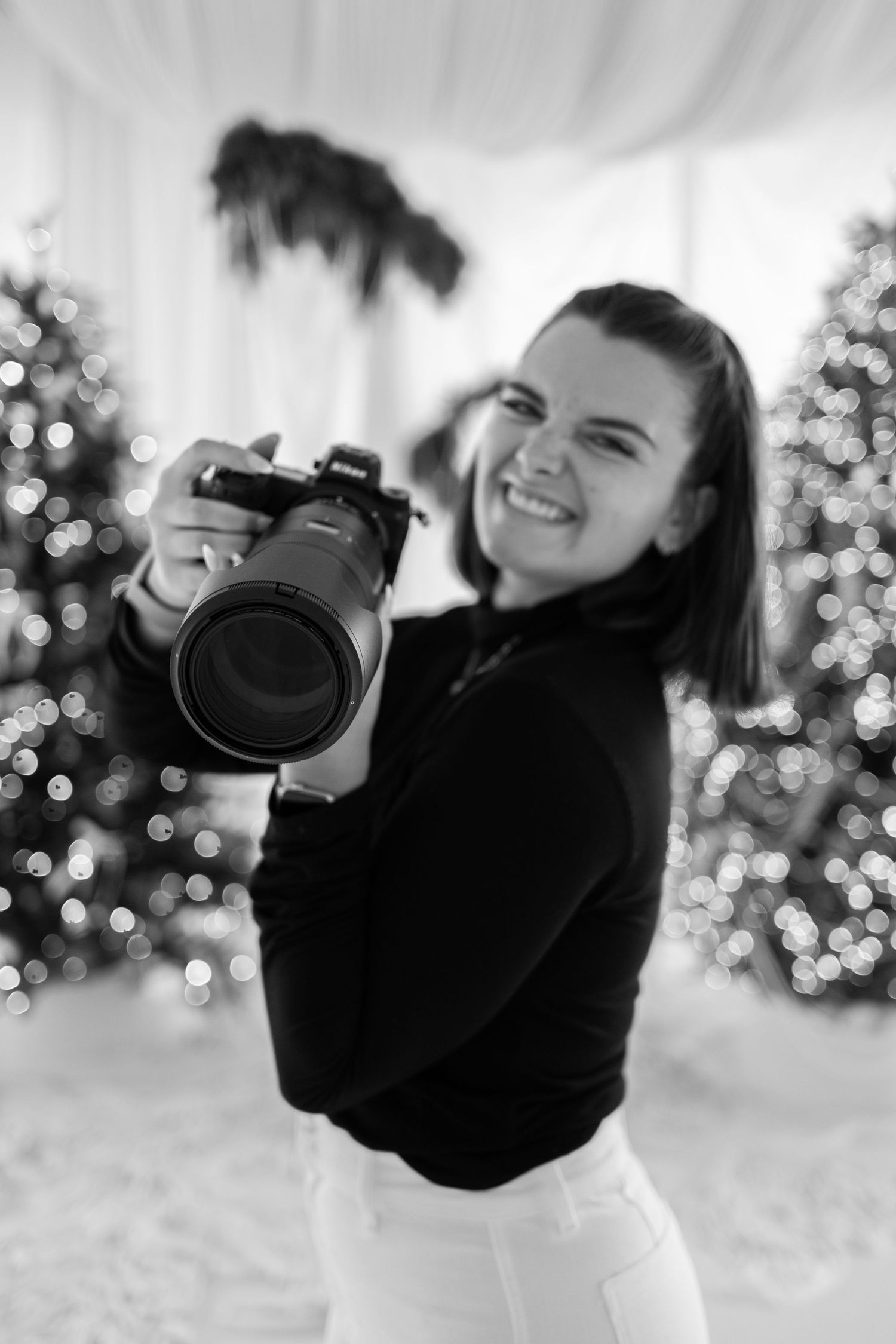 Black and white photo of a laughing female photographer holding a camera near Christmas trees.