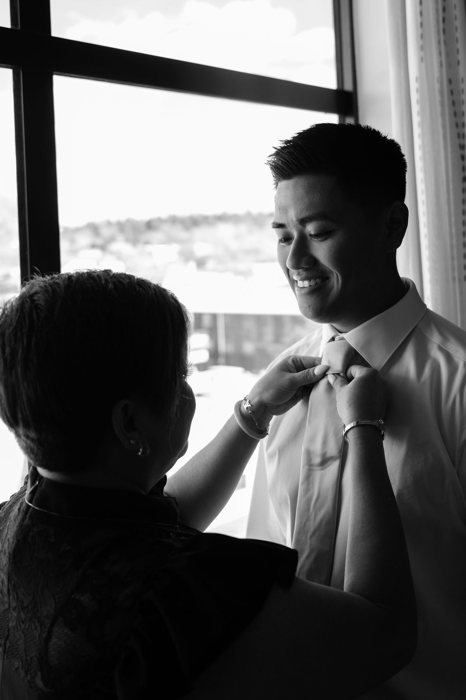 Black and white photo of smiling groom having tie adjusted by mother near bright window in Flagstaff Arizona.