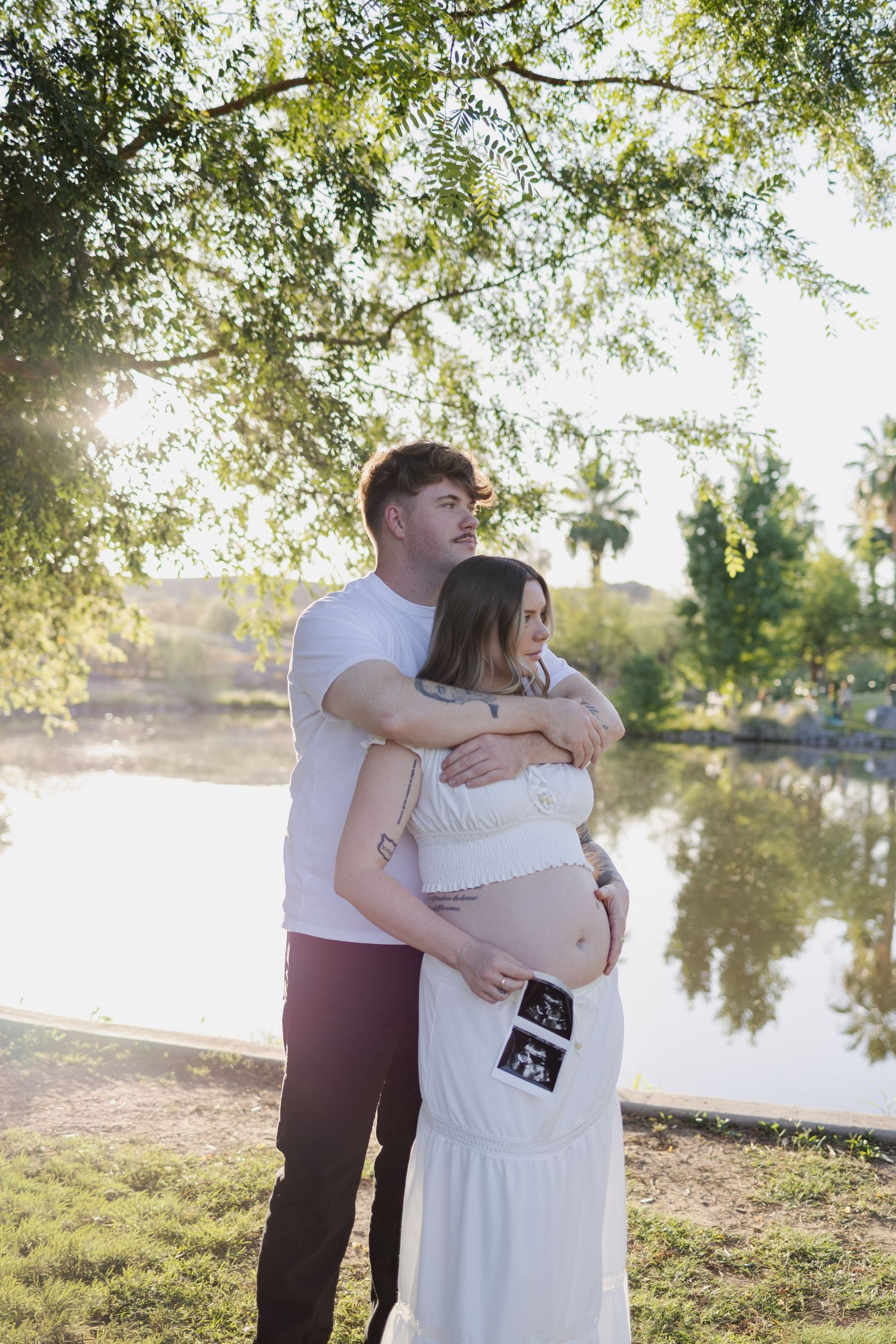 Expecting couple embracing by a sunlit lake, holding ultrasound photos against pregnant belly in a park in phoenix