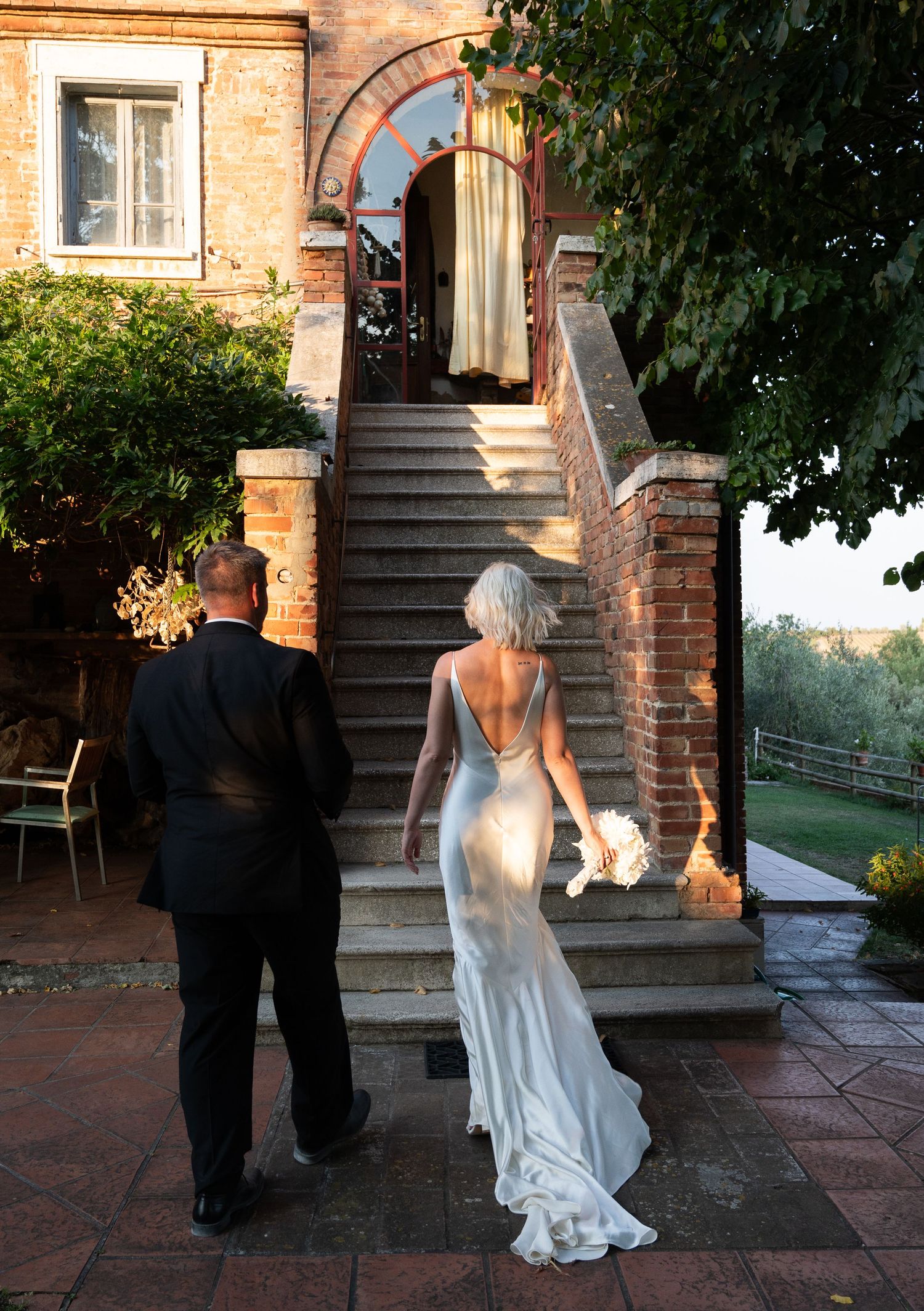 Bride in backless gown and groom ascending stone steps of a rustic Italian villa at sunset in Tuscany Italy.