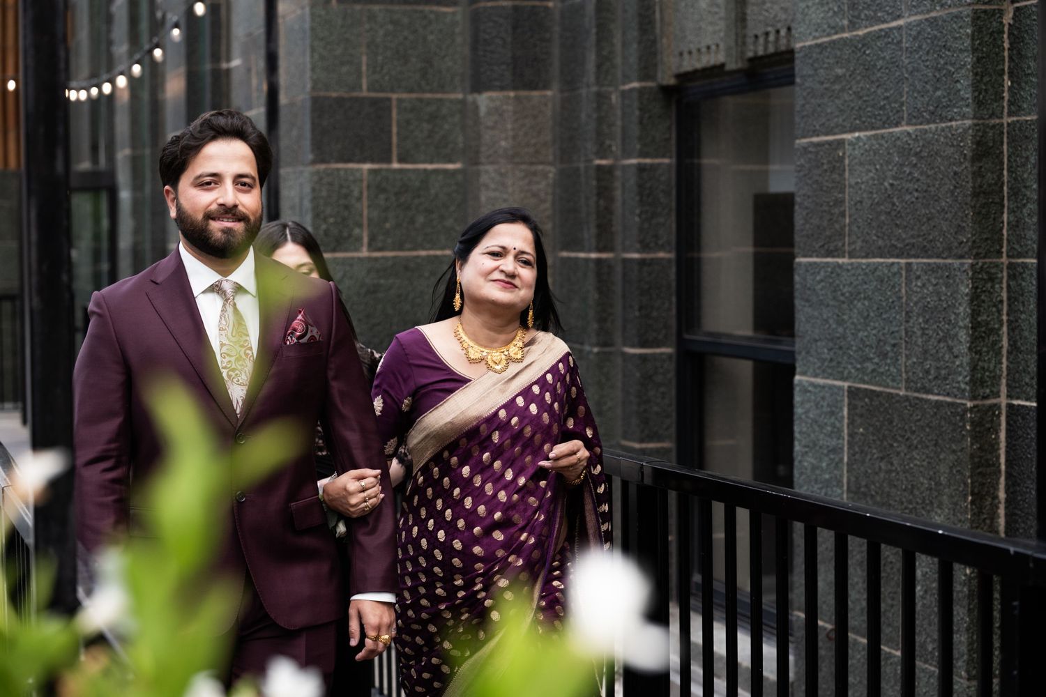 Groom in burgundy suit escorts woman in purple saree along a stone-walled venue corridor at The Pendry Hotel in Chicago Illinois.