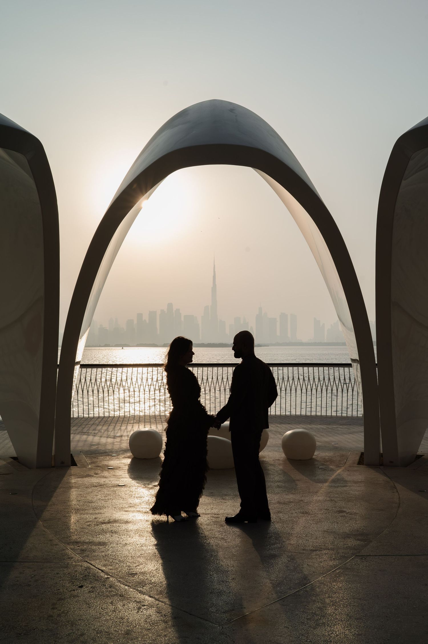 Silhouette of a couple holding hands under an arch with Dubai skyline and Burj Khalifa at sunset.