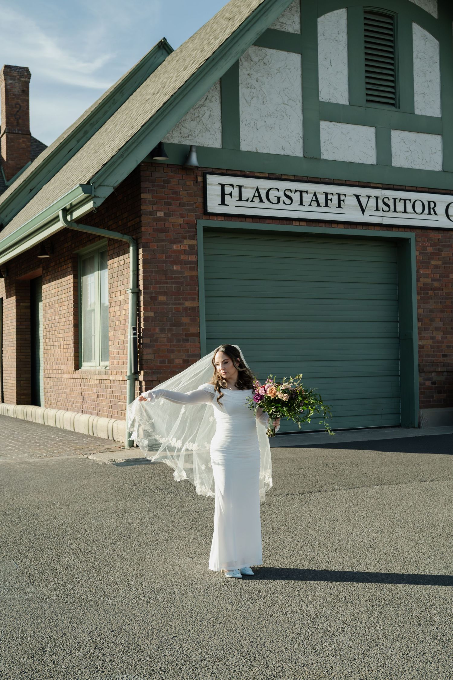 Bride in fitted white dress holds colorful bouquet outside Flagstaff Visitor Center in Flagstaff Arizona.