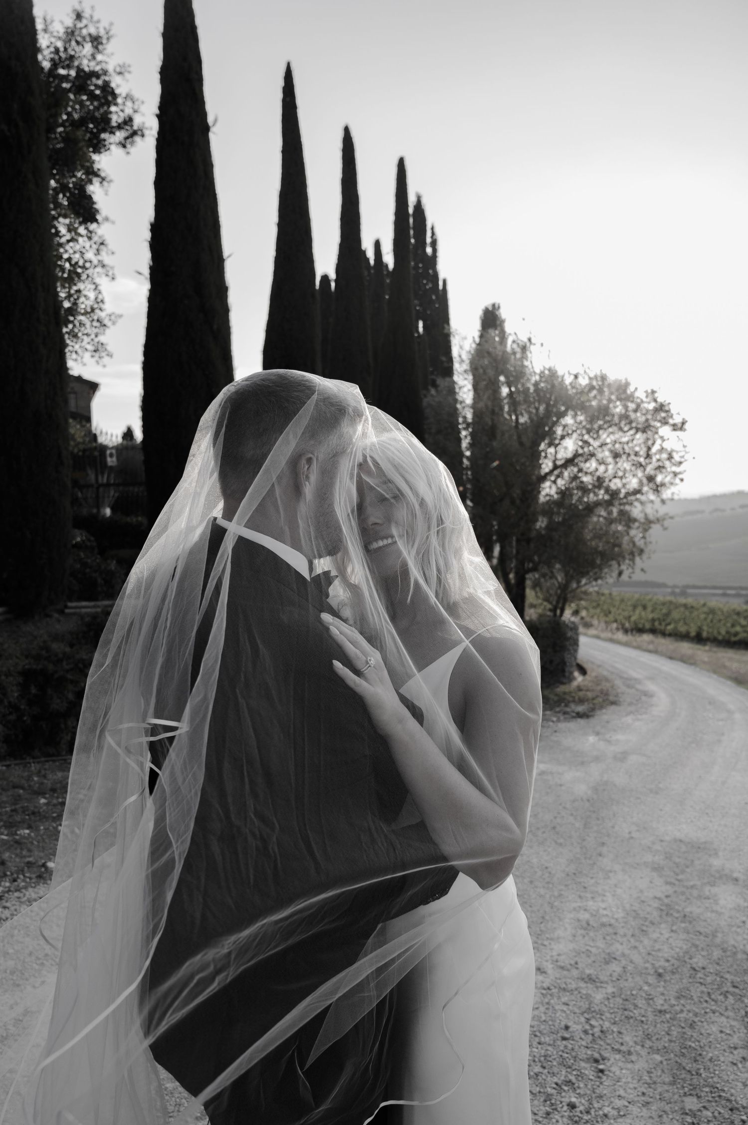 Bride and groom share a romantic kiss under a flowing wedding veil on a Tuscan cypress-lined path.