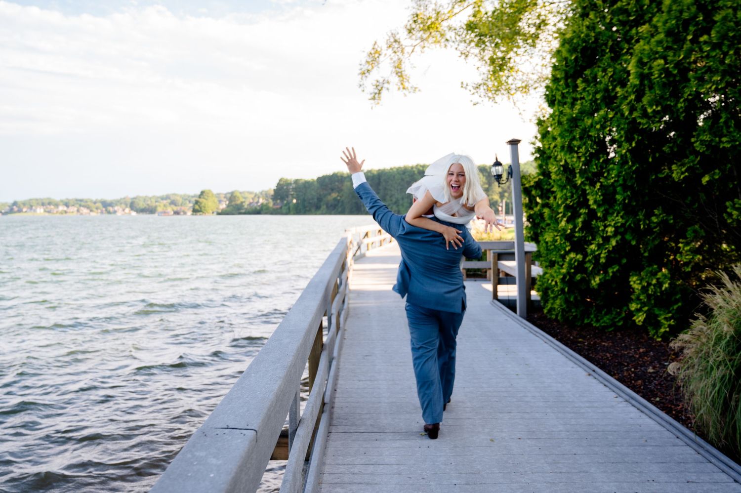 Woman with white hair waving on a waterfront boardwalk in blue outfit on a sunny day.