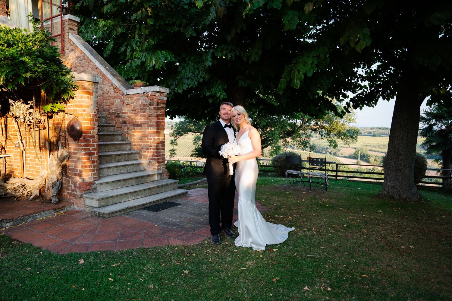 Bride and groom embrace near brick staircase on lush green lawn at outdoor wedding venue.