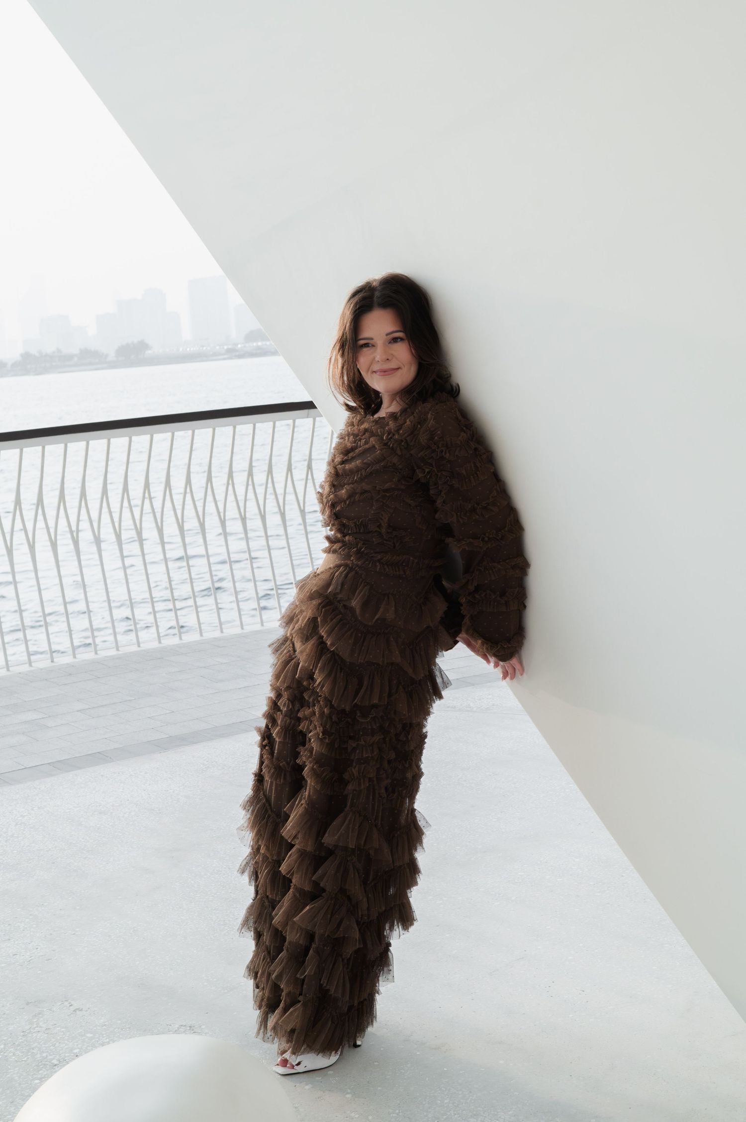 A woman in a textured brown ruffled jumpsuit leans against a white wall with a waterfront view behind her.