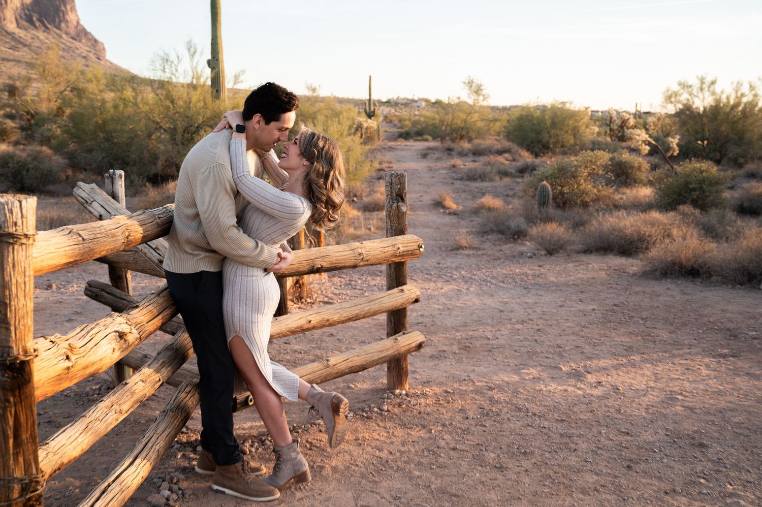 Couple sharing a romantic kiss leaning against a rustic wooden fence in a desert landscape at golden hour.
