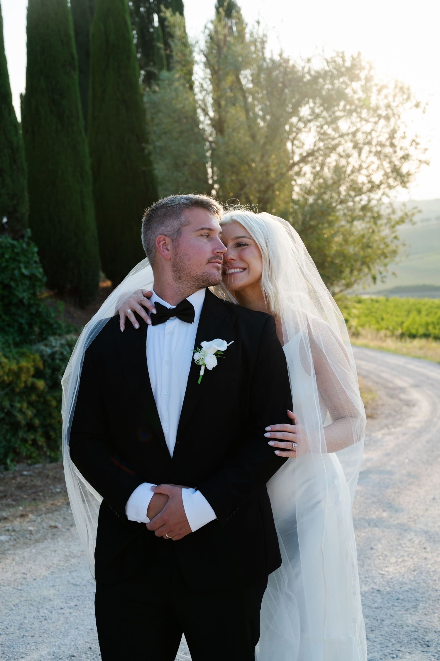 Groom in black tuxedo kissing bride in white veil at golden hour in a vineyard setting in Tuscany Italy.