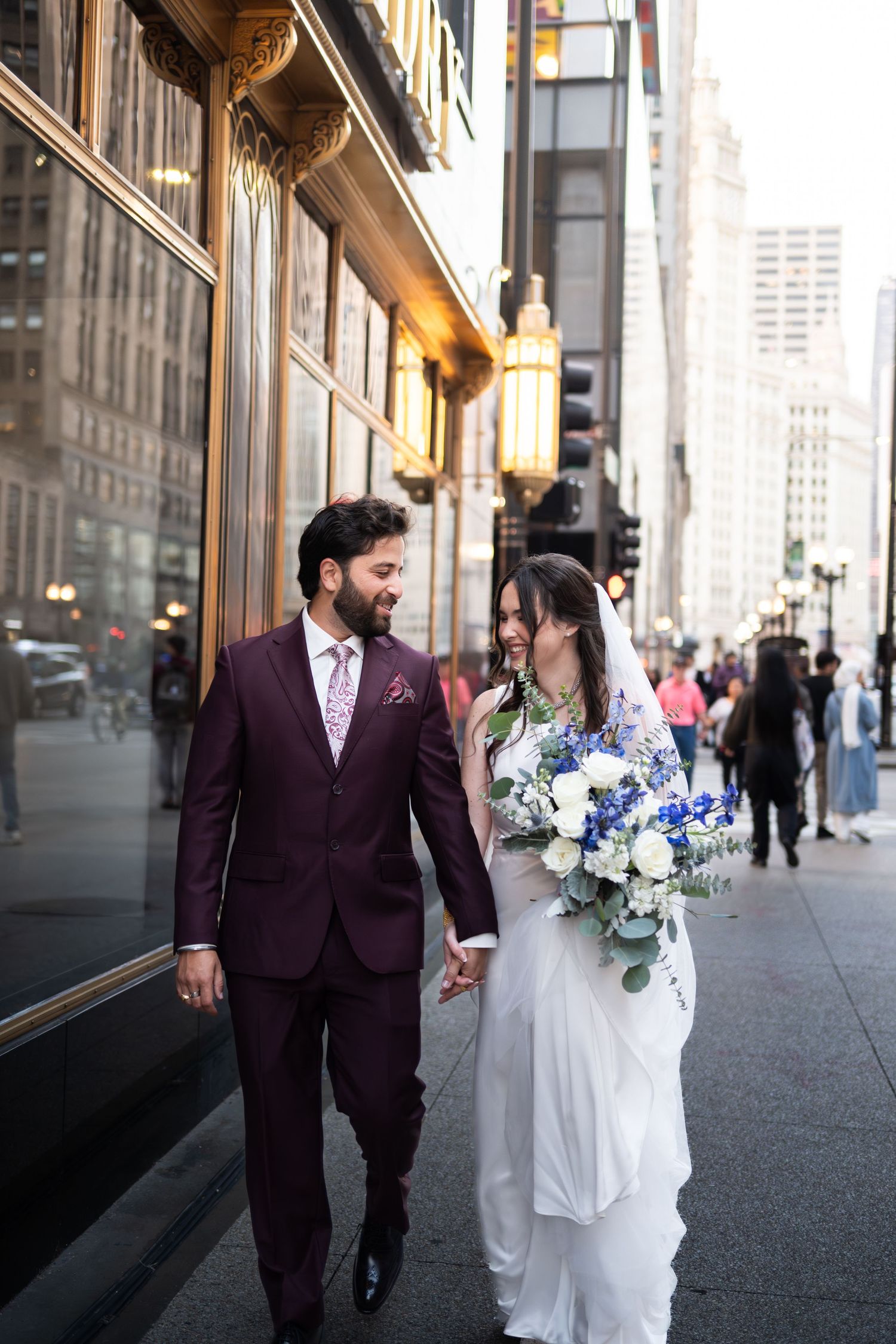 Bride and groom walk hand-in-hand on a city street, she holds blue and white bouquet, he wears a burgundy suit In Chicago