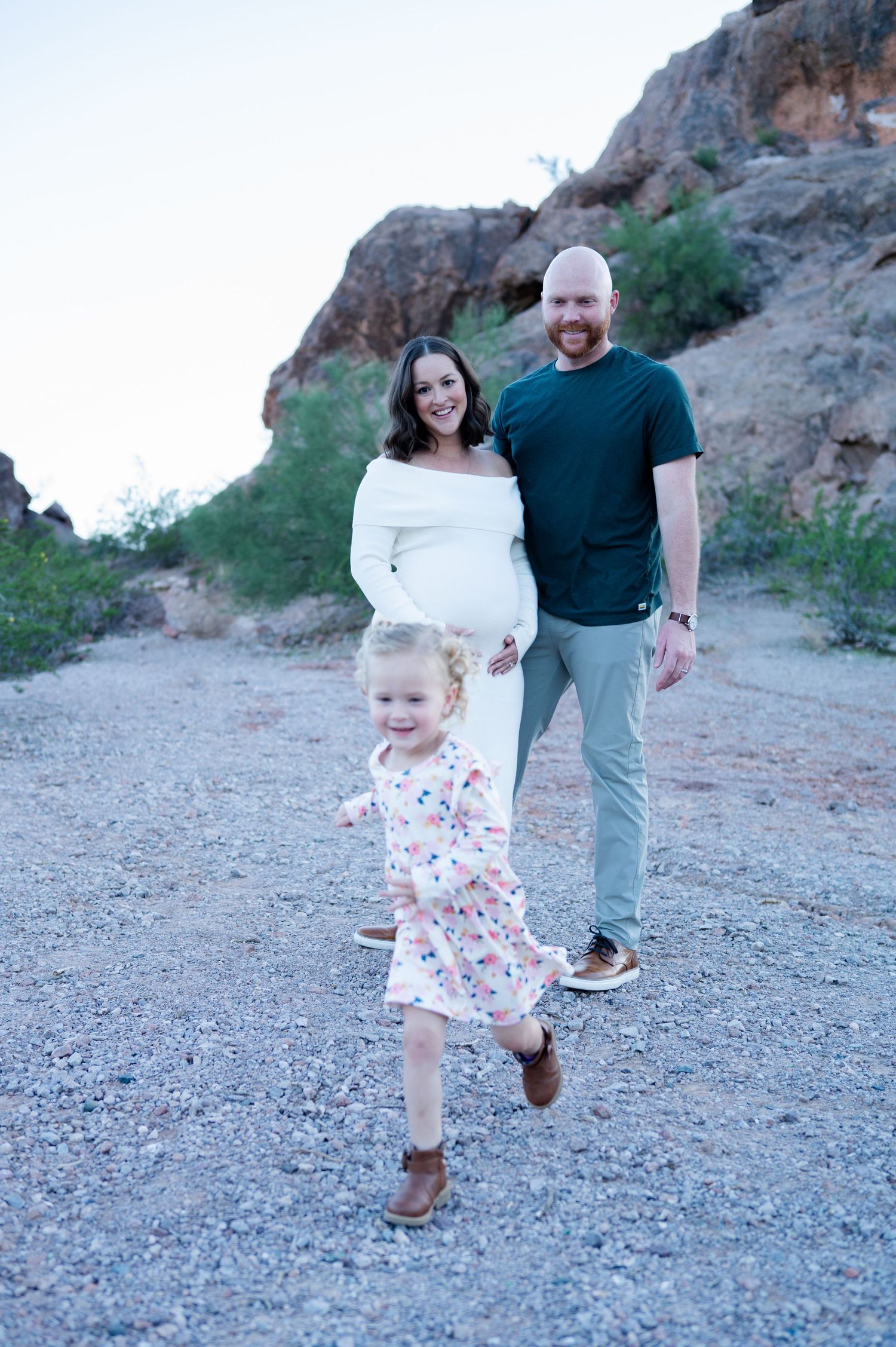 Pregnant woman in white dress poses with husband and toddler daughter on desert trail with rocky landscape.