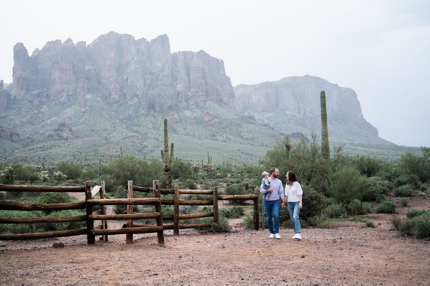 Couple standing by a rustic fence with Superstition Mountains and saguaro cacti in the Arizona desert background. Lost Dutchman State Park