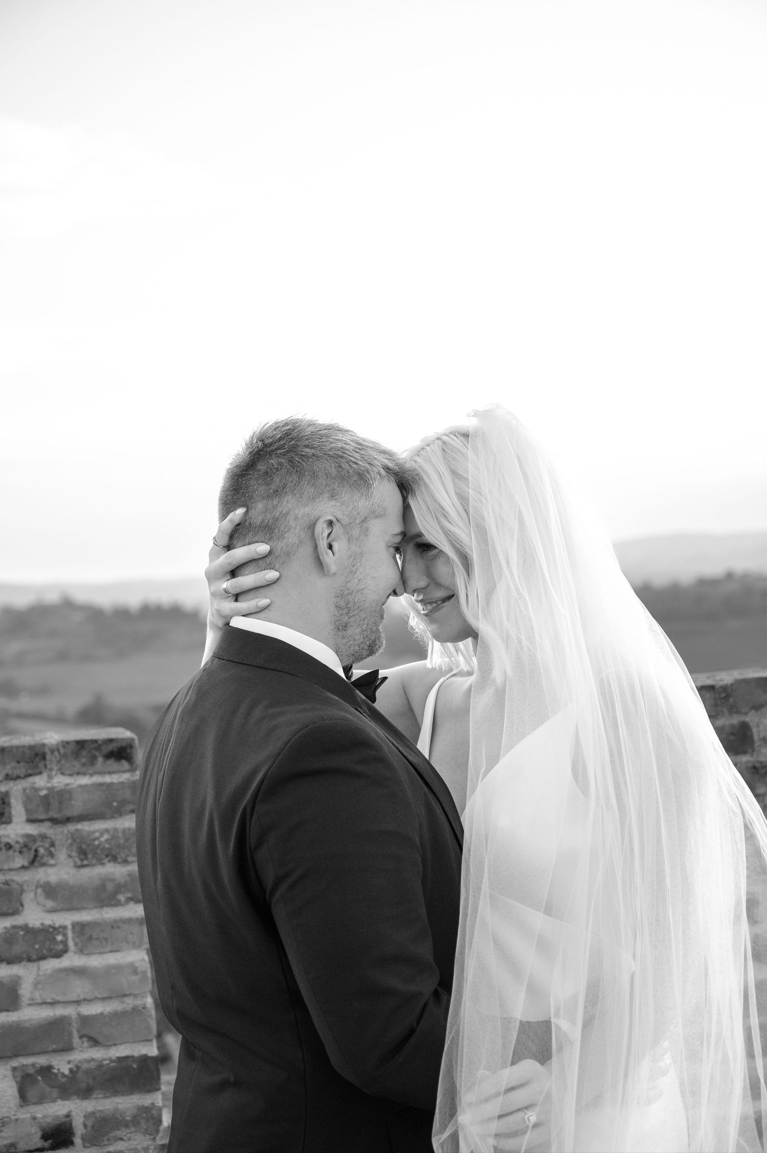 Black and white portrait of bride and groom sharing intimate moment under veil at rustic brick wall in Tuscany Italy.