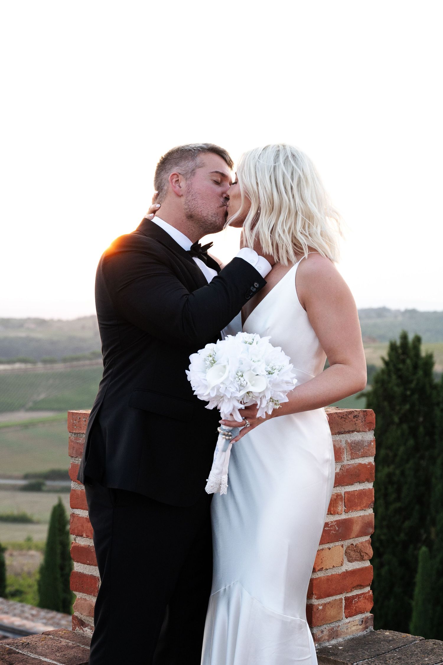 A bride and groom share a romantic kiss outdoors, she holds white bouquet, wearing wedding dress, he in black tuxedo.