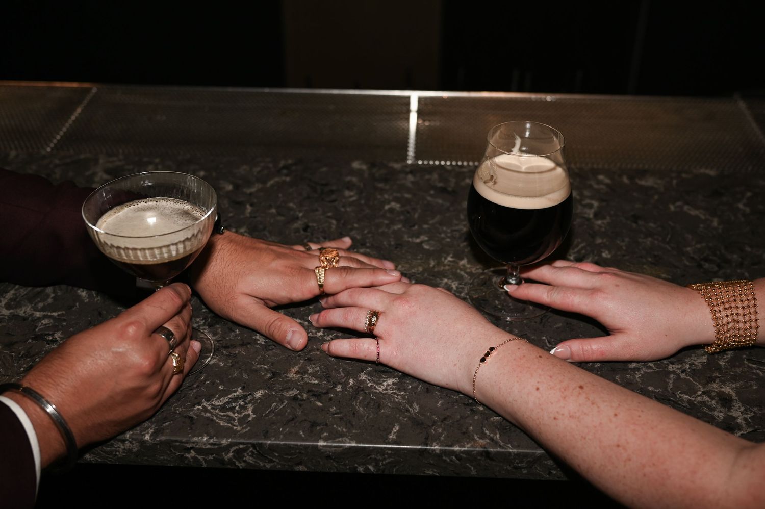 Two people hold hands on a marble bar top beside dark cocktails at a wedding celebration at The Pendry Hotel in Chicago Illinois.
