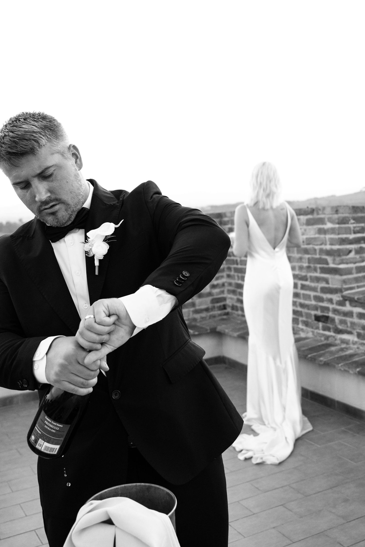 Groom in black tuxedo opening champagne bottle on rooftop terrace as bride stands by brick wall in Tuscany Italy.
