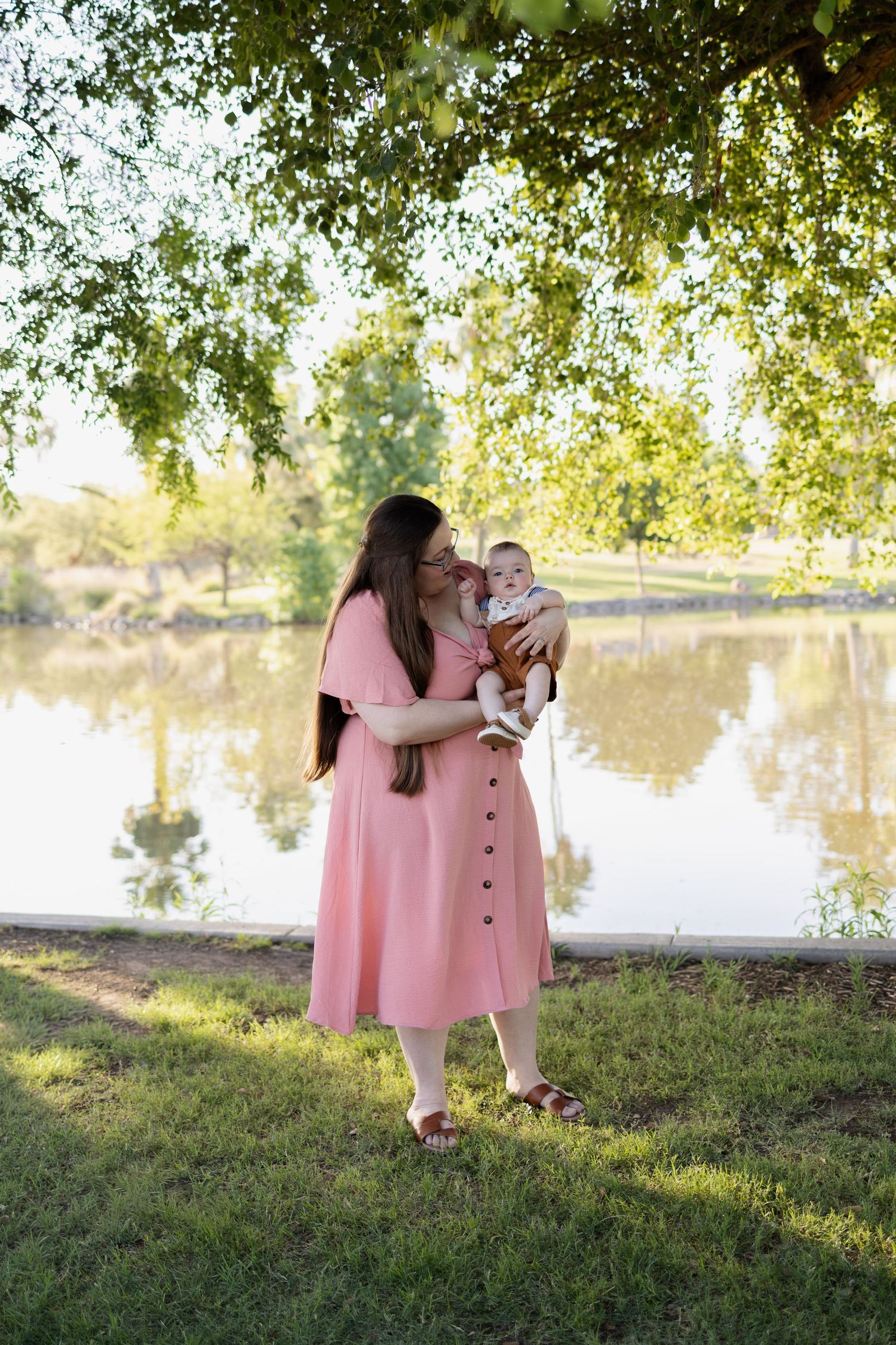A mother in a pink dress holds and kisses her baby by a reflective pond under green trees.
