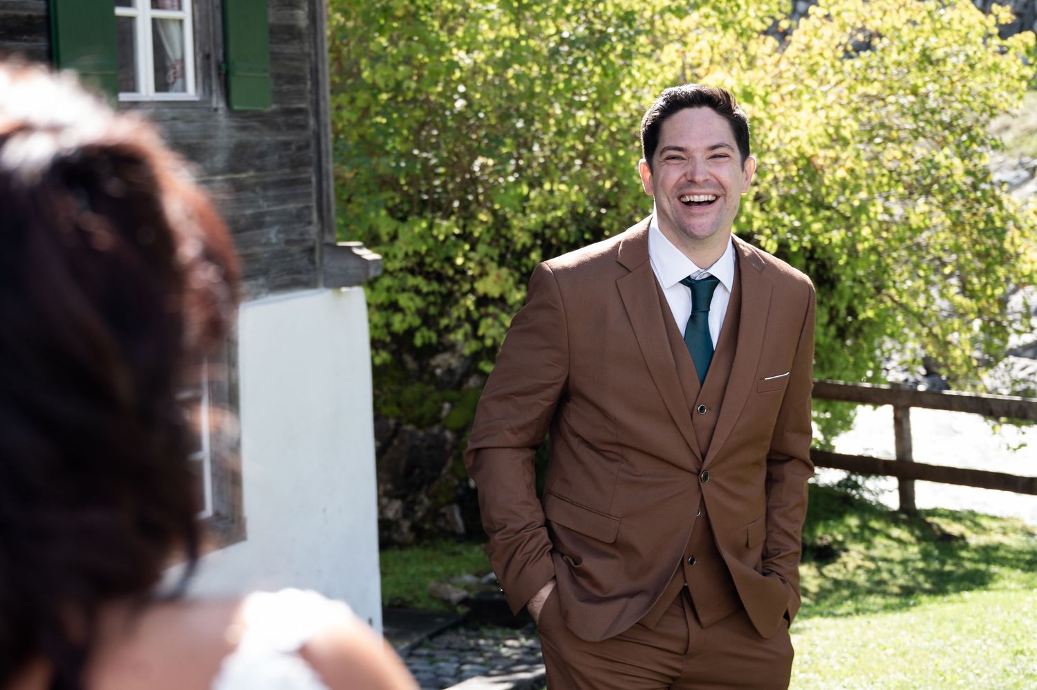 Joyful groom in brown suit laughing during outdoor wedding ceremony in a sunny garden setting in Switzerland.