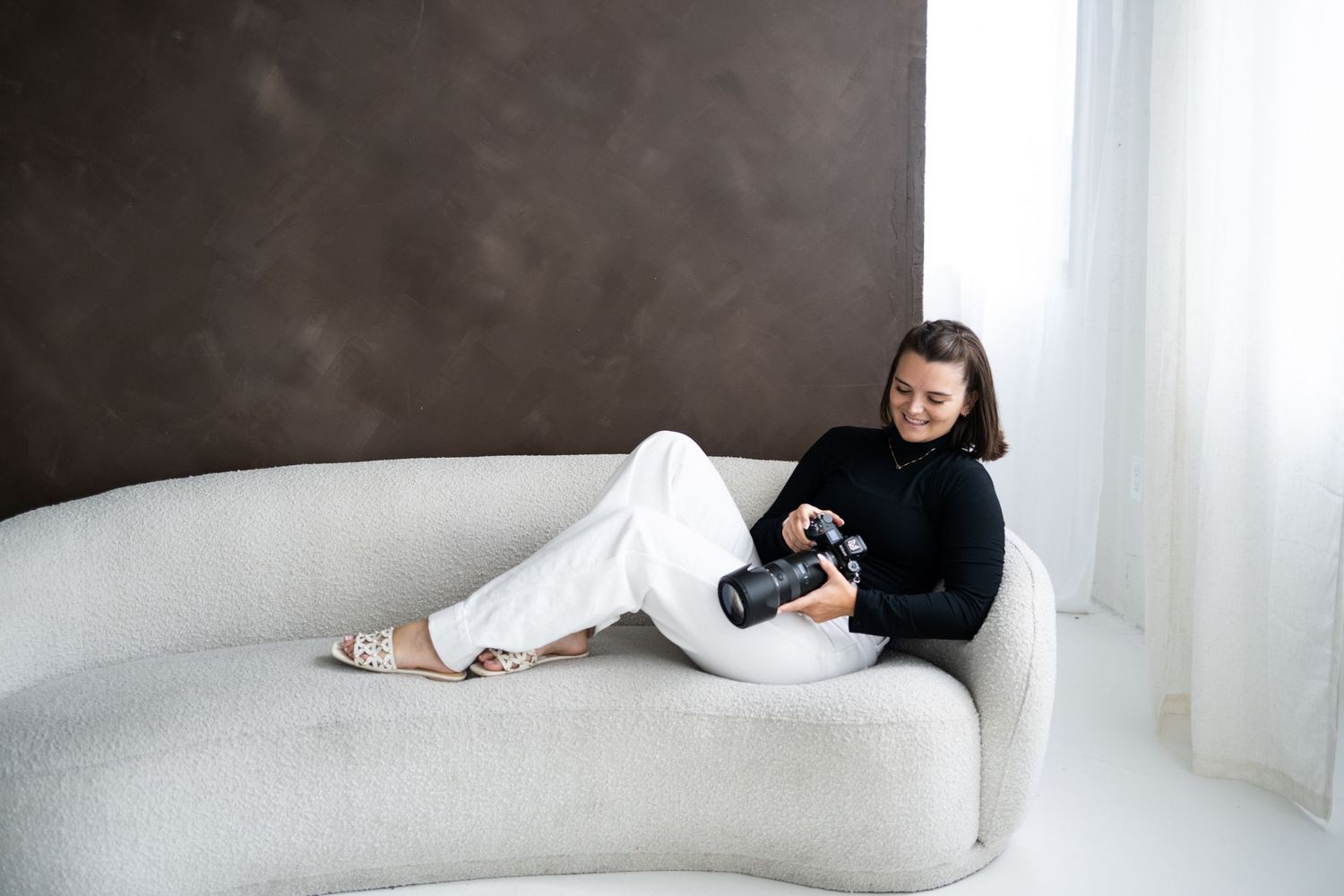 Female photographer sits on a white curved sofa reviewing photos on her Nikon DSLR camera indoors.