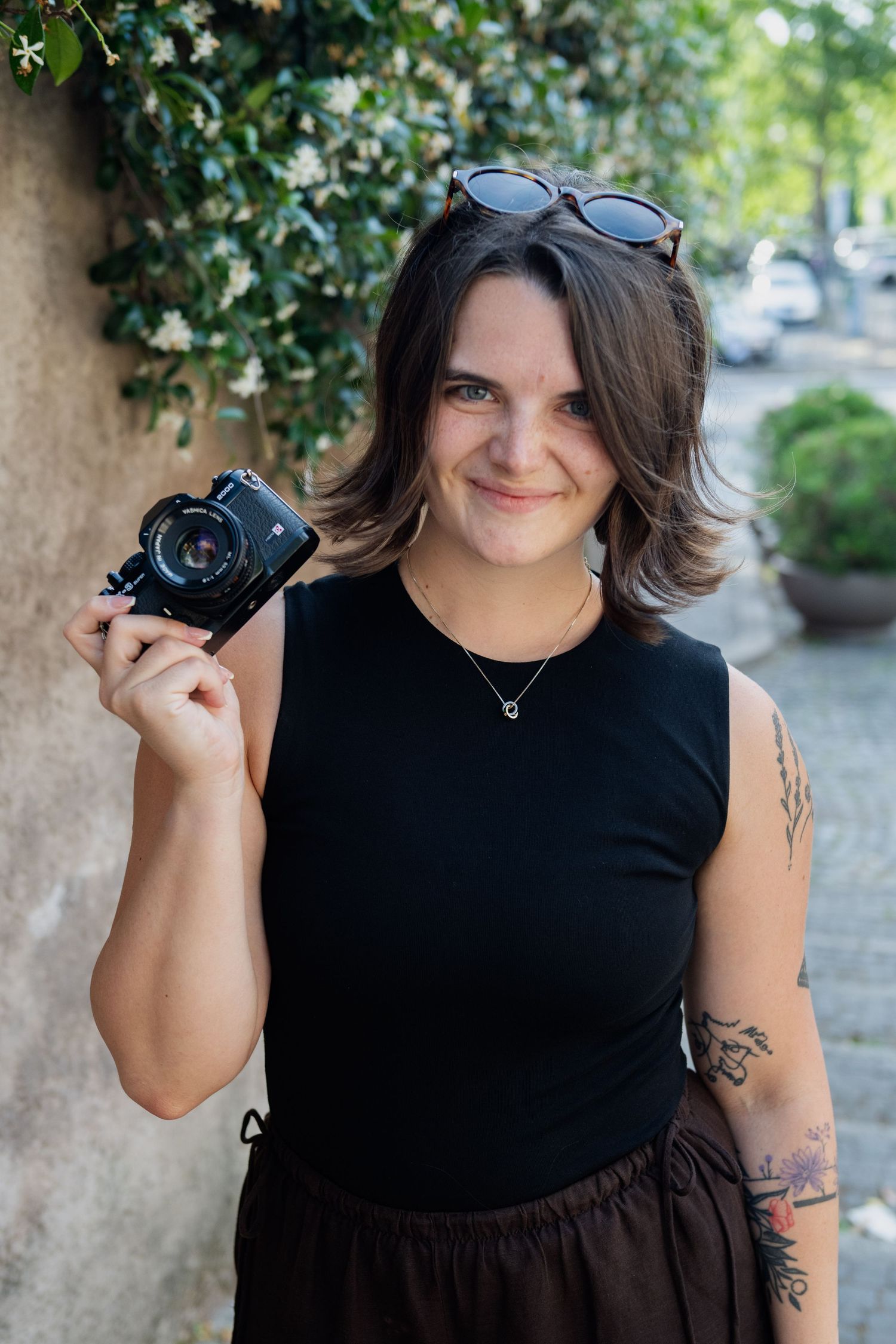 Smiling female photographer holds a vintage film camera outdoors with sunglasses on her head.