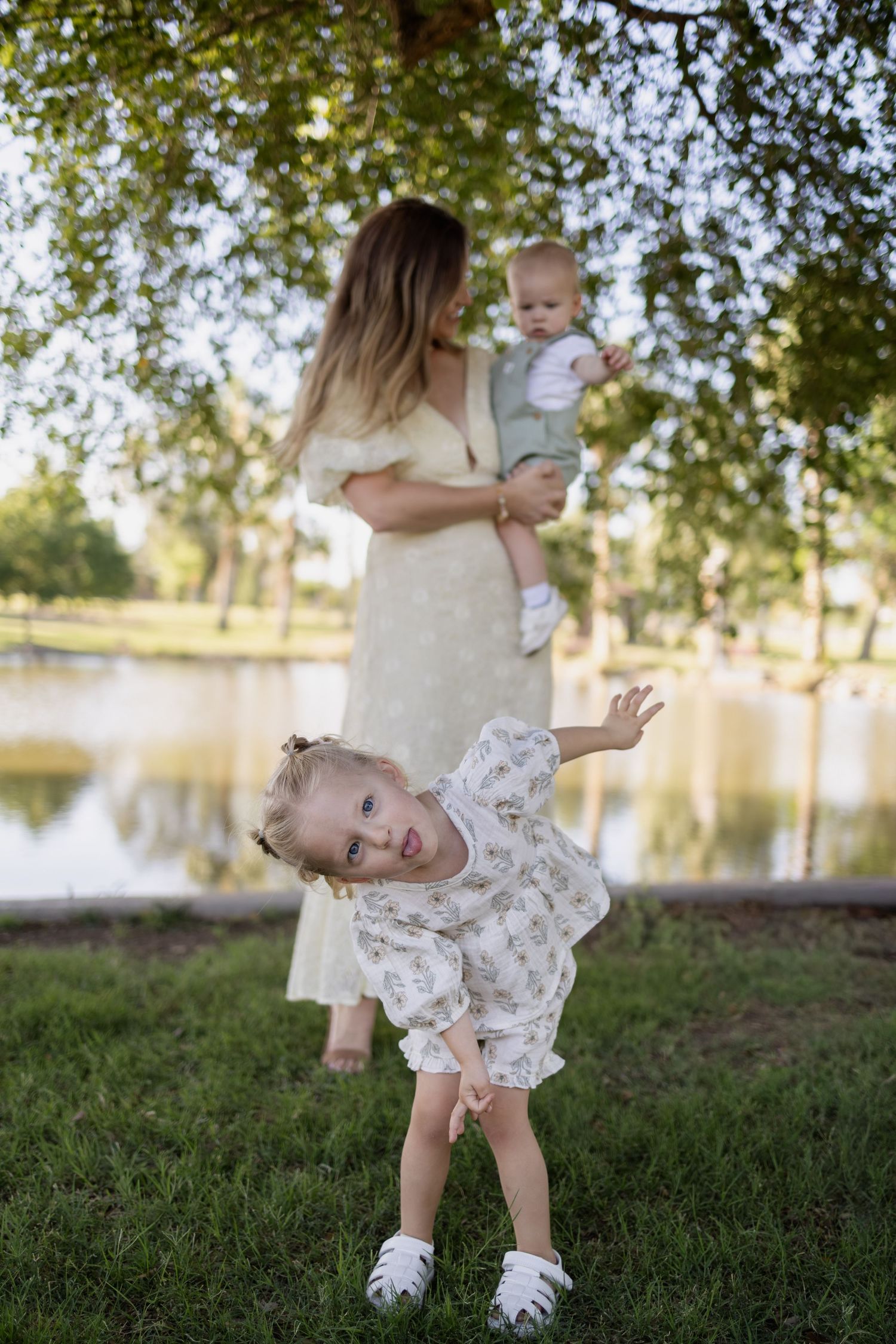 Mother in yellow dress holds baby while toddler girl plays outdoors near a pond in a lush green park.