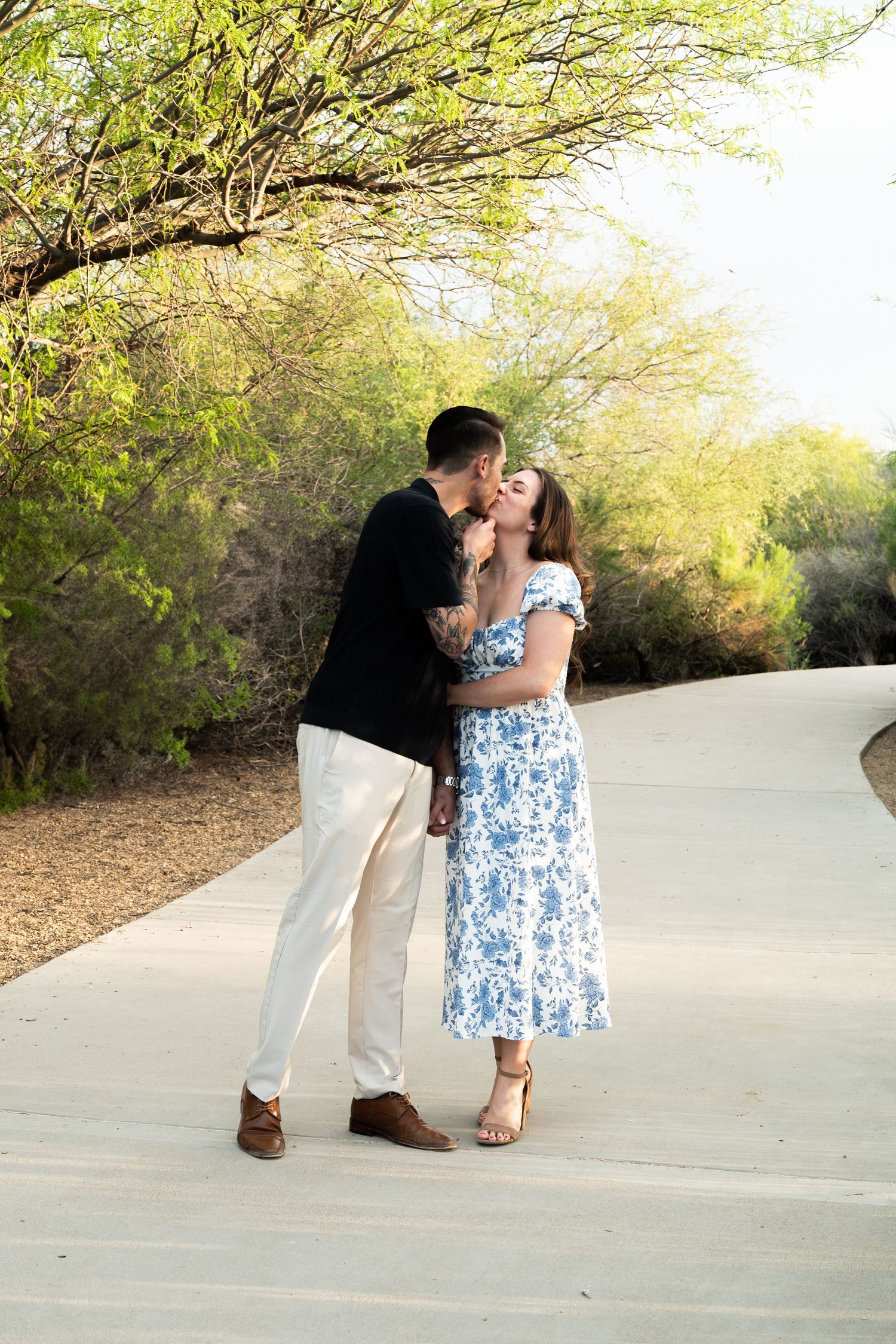 Couple sharing a kiss on a sunlit pathway surrounded by lush green desert trees and shrubs In arizona.