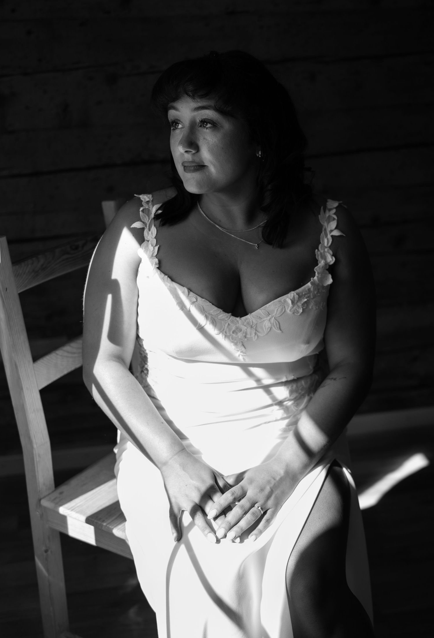 Bride in white slip dress seated on a wooden chair in dramatic black and white portrait lighting in Lauterbrunnen Switzerland.