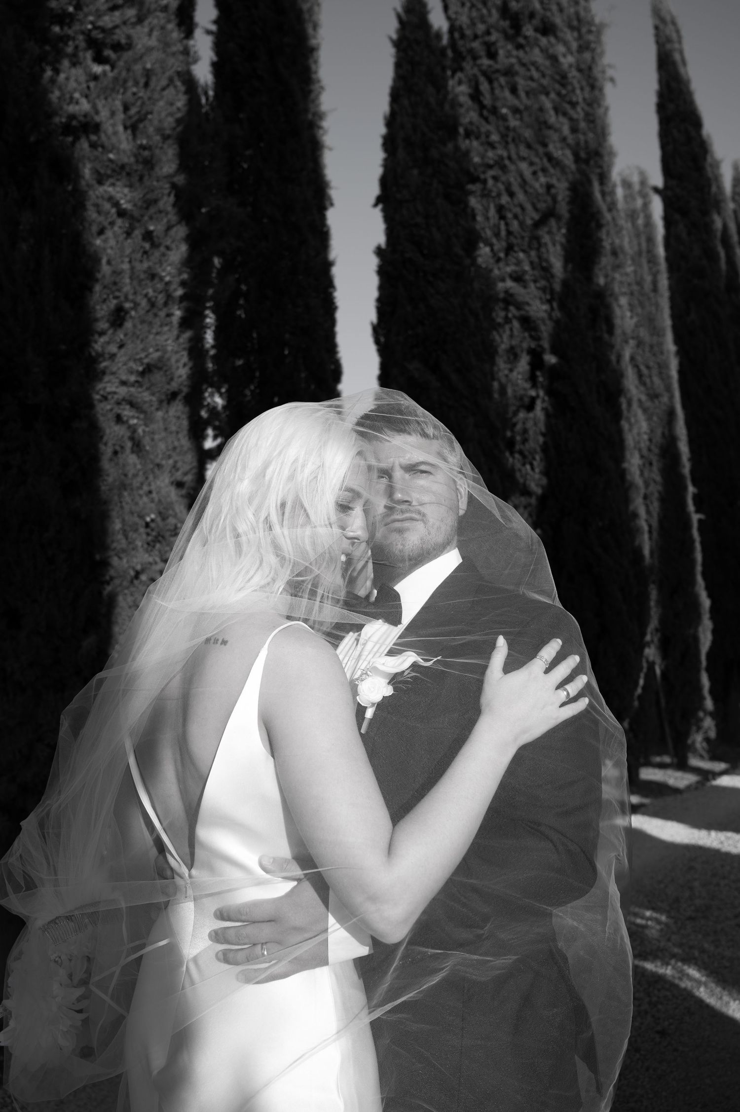 Black and white romantic portrait of bride and groom embracing under veil among tall cypress trees in Tuscany Italy.