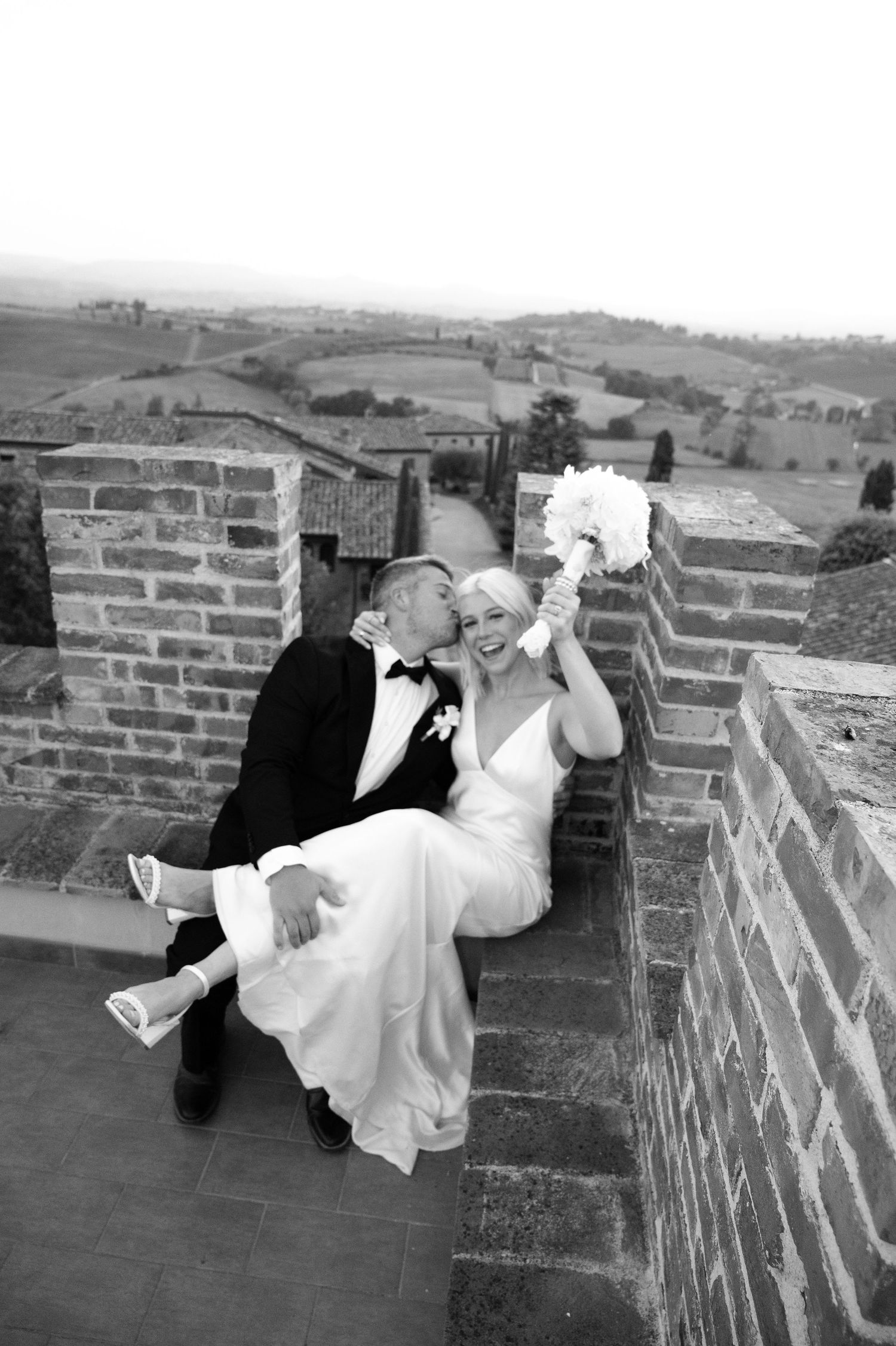 Black and white photo of groom kissing bride on cheek, sitting on Tuscan villa terrace, bride holding bouquet.