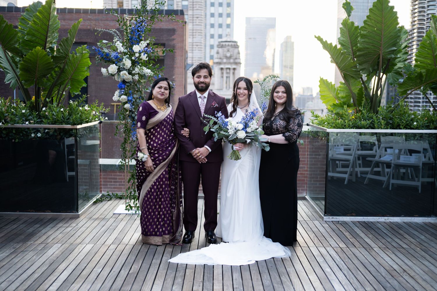 Wedding party poses on rooftop terrace, bride in white gown with bouquet, groom in burgundy suit, guests in formal attire.