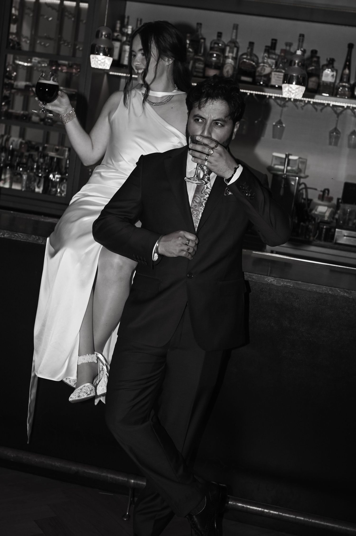 Black and white photo of bride sitting on bar beside groom holding wine glass at reception in Chicago