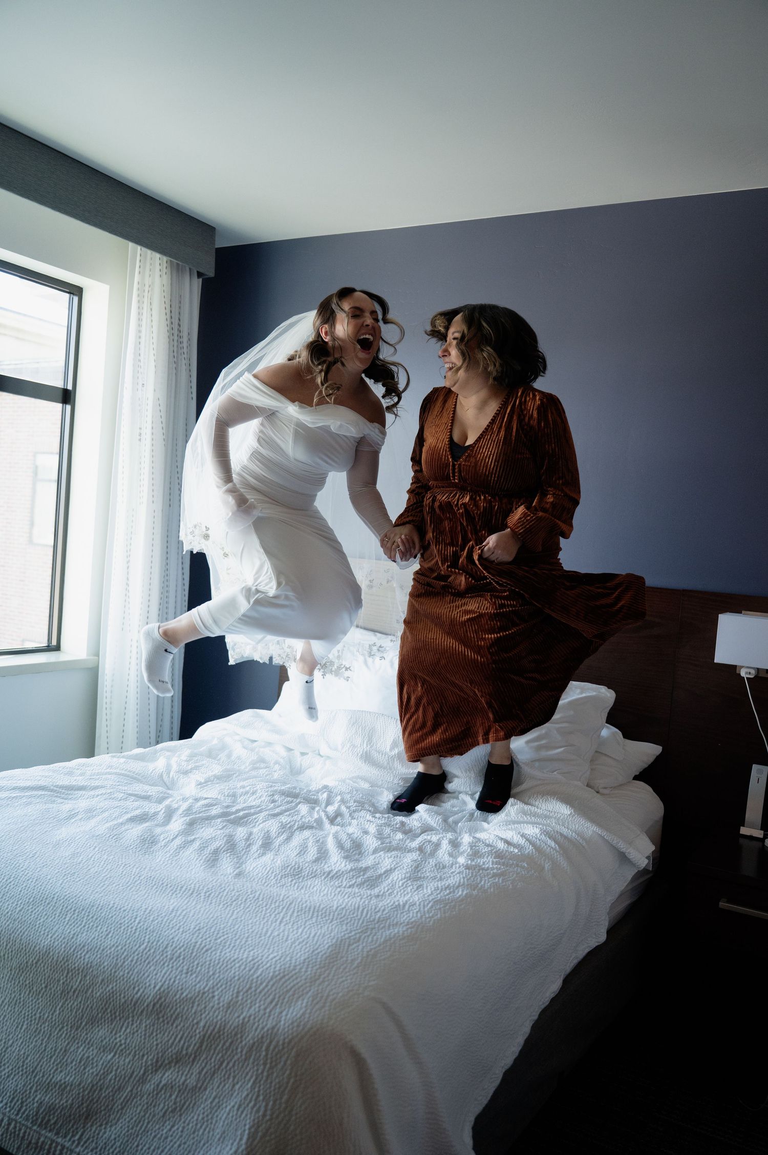 Excited bride in white gown and veil jumping on hotel bed with bridesmaid in rust dress in Flagstaff Arizona.