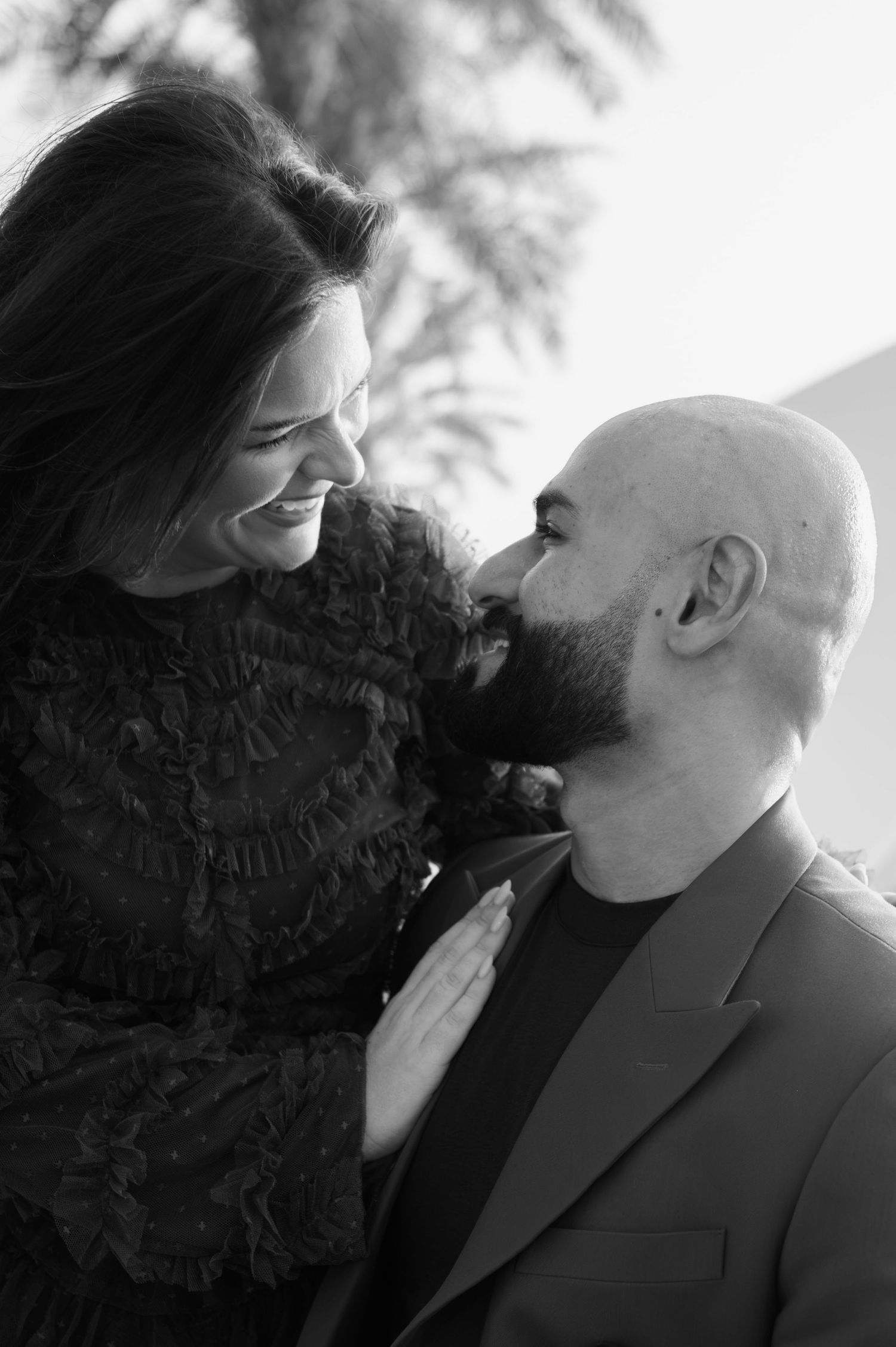 Black and white photo of a smiling woman and bearded bald man gazing at each other romantically outdoors.