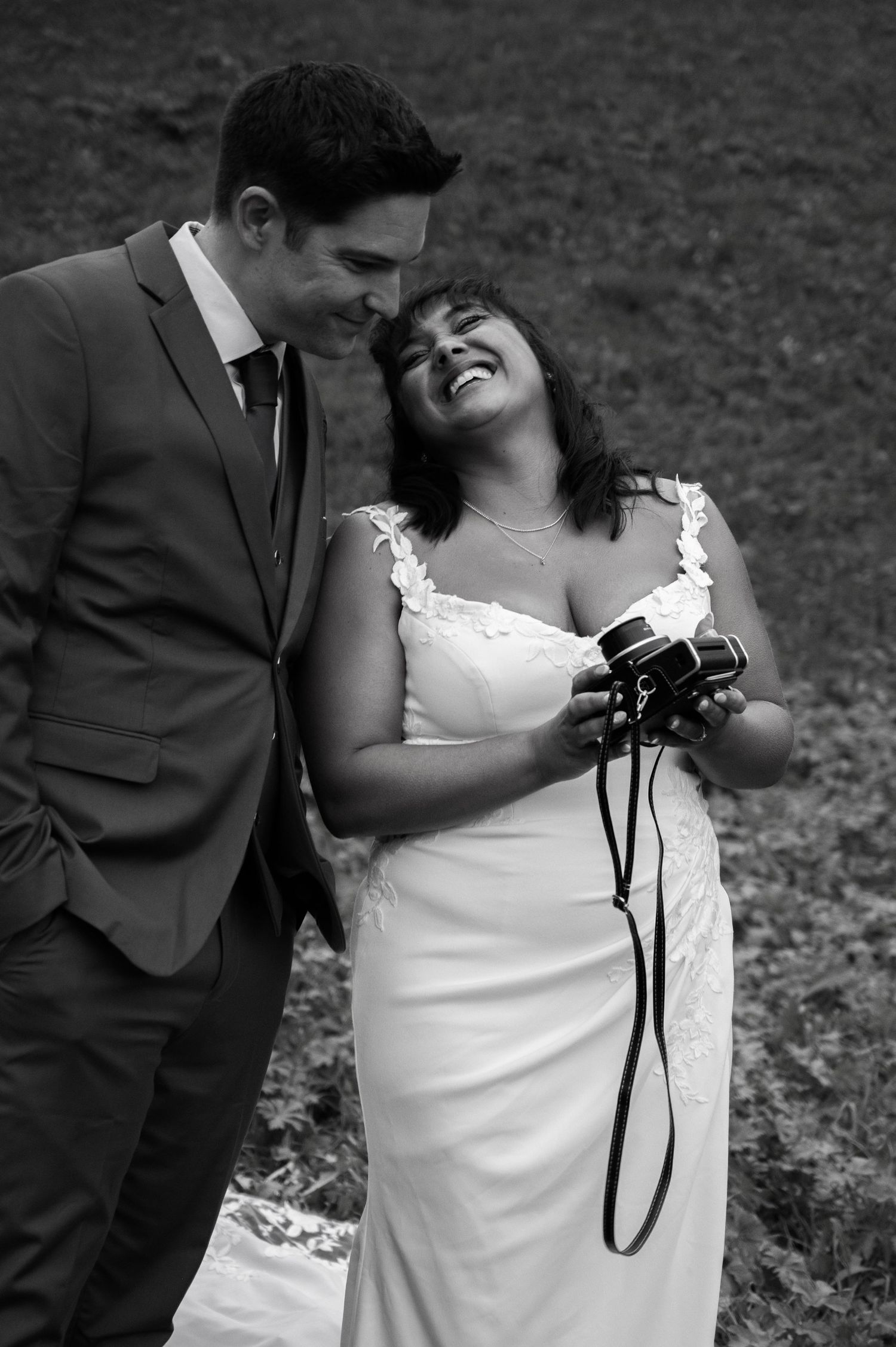 Bride laughing joyfully holding vintage camera while groom smiles at her in black and white wedding portrait in Switzerland.
