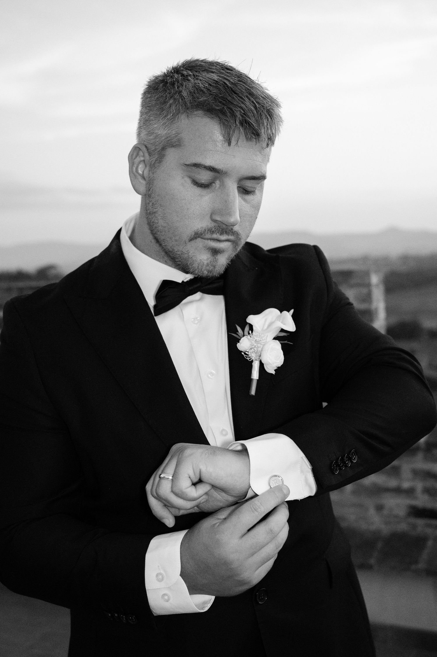 Dapper groom in black tuxedo adjusting cufflinks with boutonniere in black and white wedding portrait in Tuscany Italy.