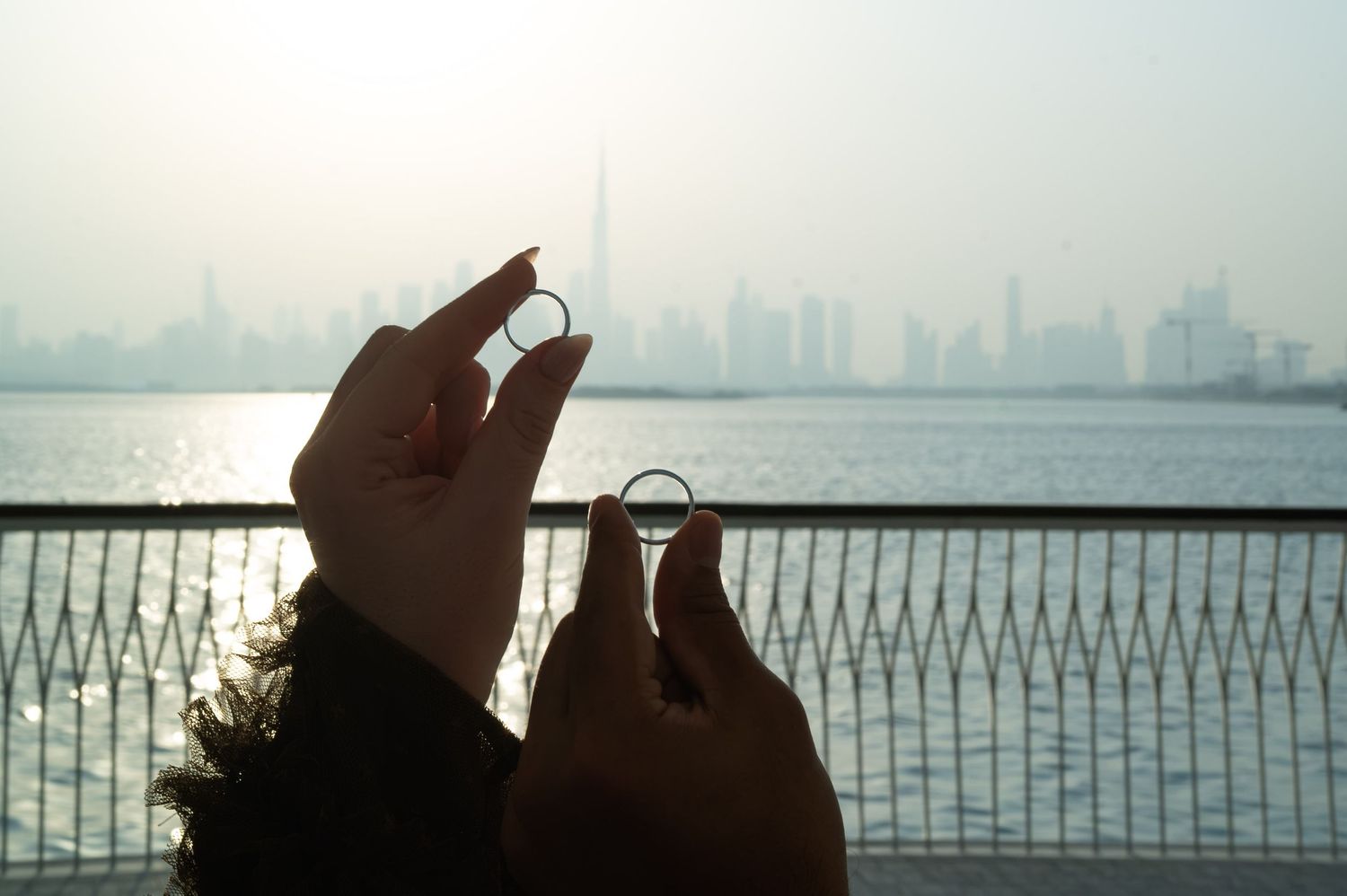 Two hands holding rings against a hazy Dubai skyline and waterfront at sunset.