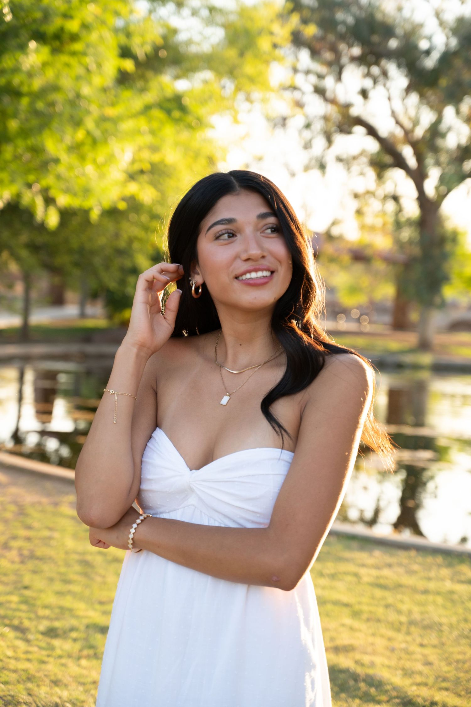 Young woman in white strapless dress smiling outdoors near a pond in a sunny park setting.